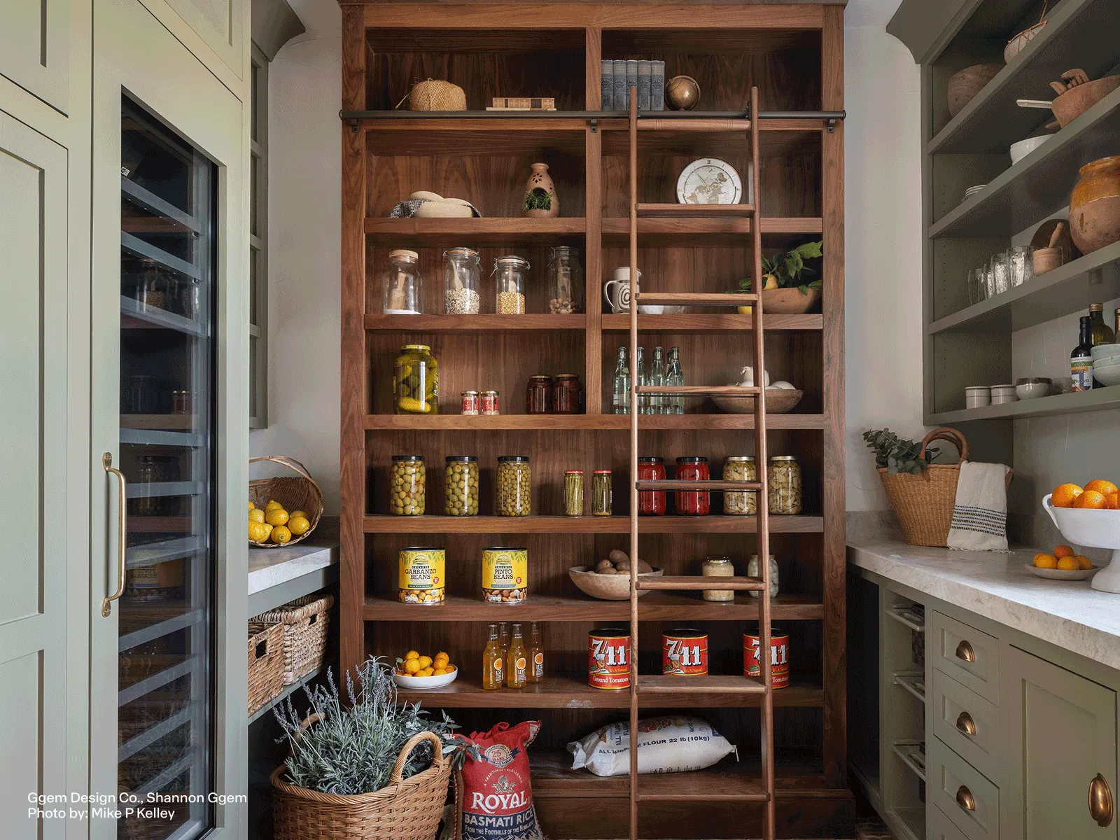 Spacious pantry with wooden shelves, jars of preserved foods, baskets, a sliding ladder, and adjacent cabinetry with countertop and fruit bowls.