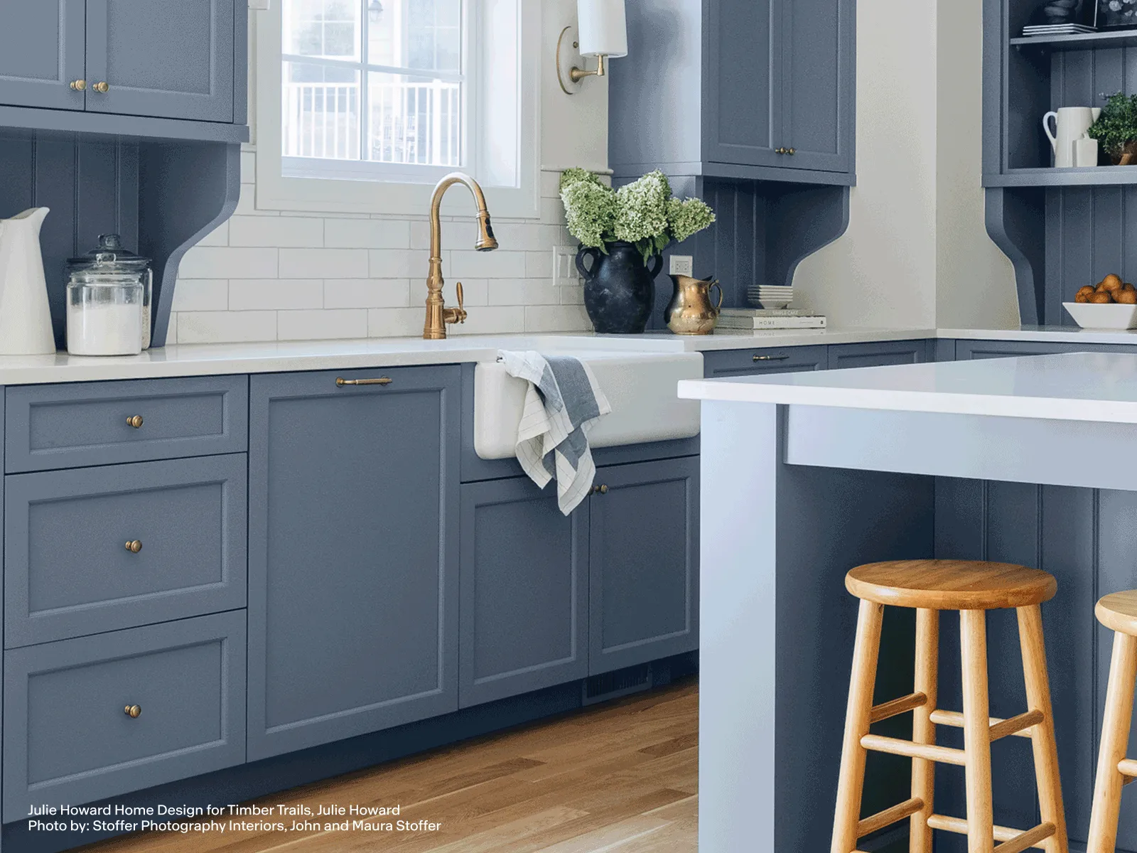 Modern kitchen with blue-gray cabinets, white countertops, wooden stools, gold faucet, and a vase of greenery by a large window.