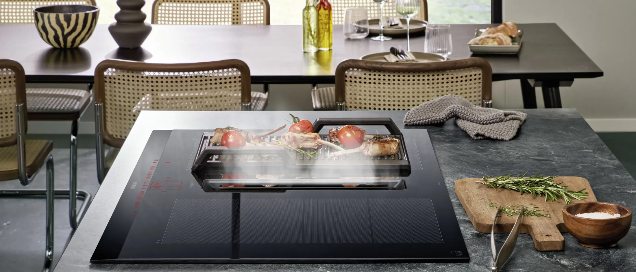 Modern kitchen island with an induction grill cooking meat and tomatoes, surrounded by dining table and chairs with tableware and herbs.