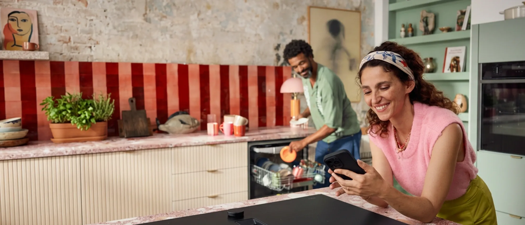 Lächelnde Frau in einem rosa Pullover, die ein Smartphone benutzt, während ein Mann in einer gemütlichen Küche einen Geschirrspüler belädt.