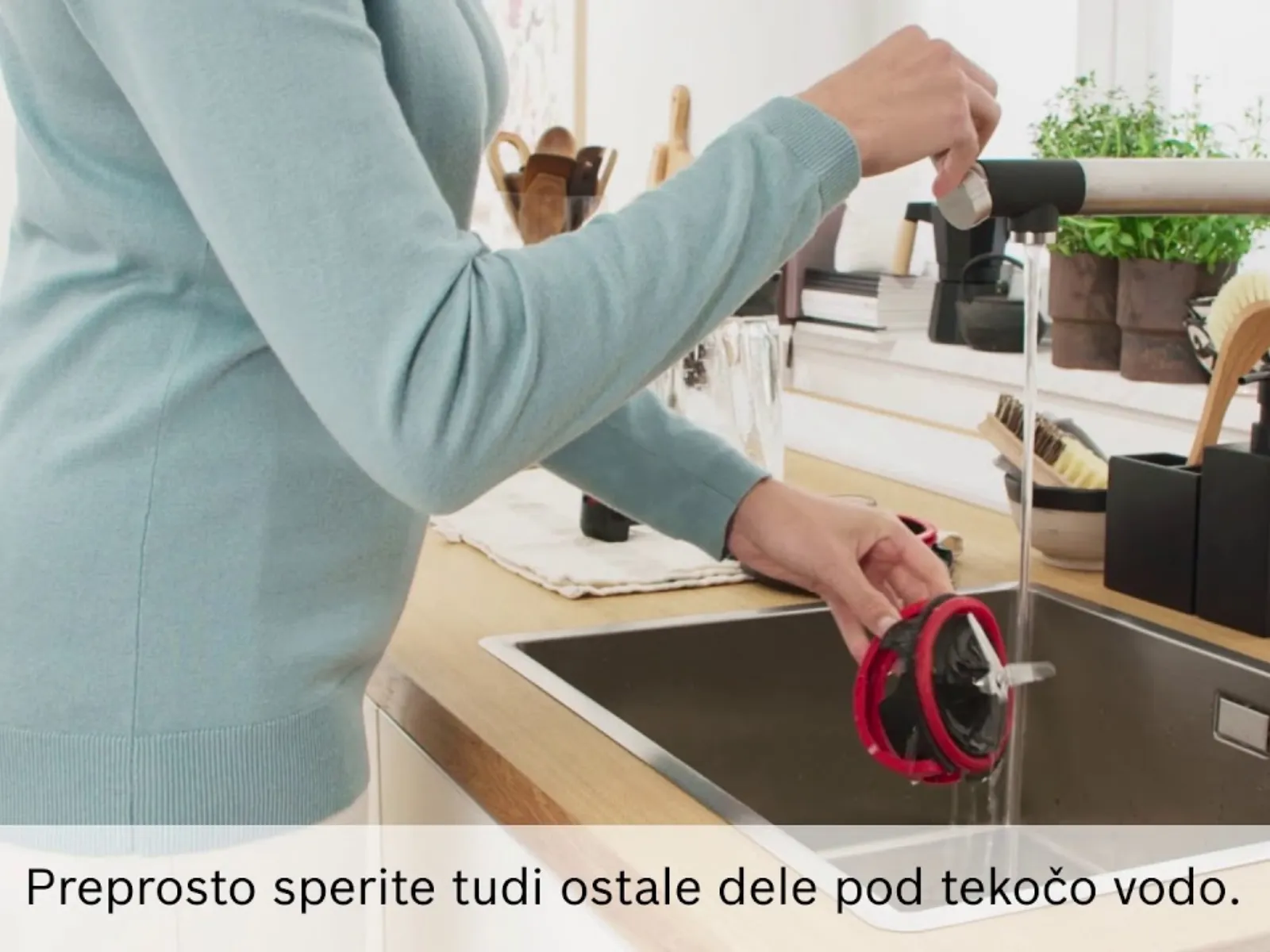 Oseba, ki pod tekočo vodo v pomivalnem koritu spira rdeč in črn del kuhinjskega aparata