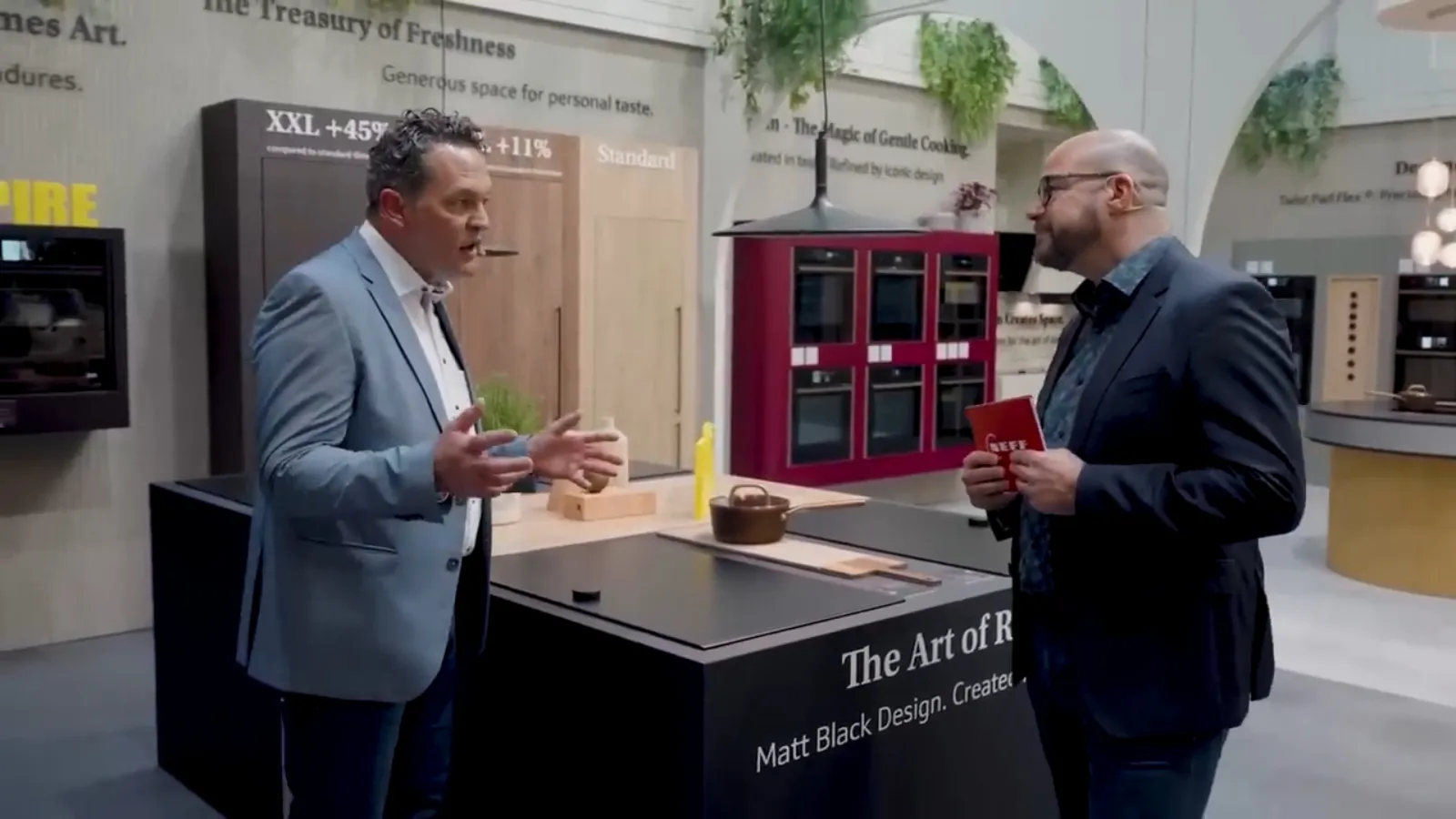 Two men discuss kitchen appliances in a modern NEFF showroom, standing by a display cooktop with red built-in ovens visible in the background.