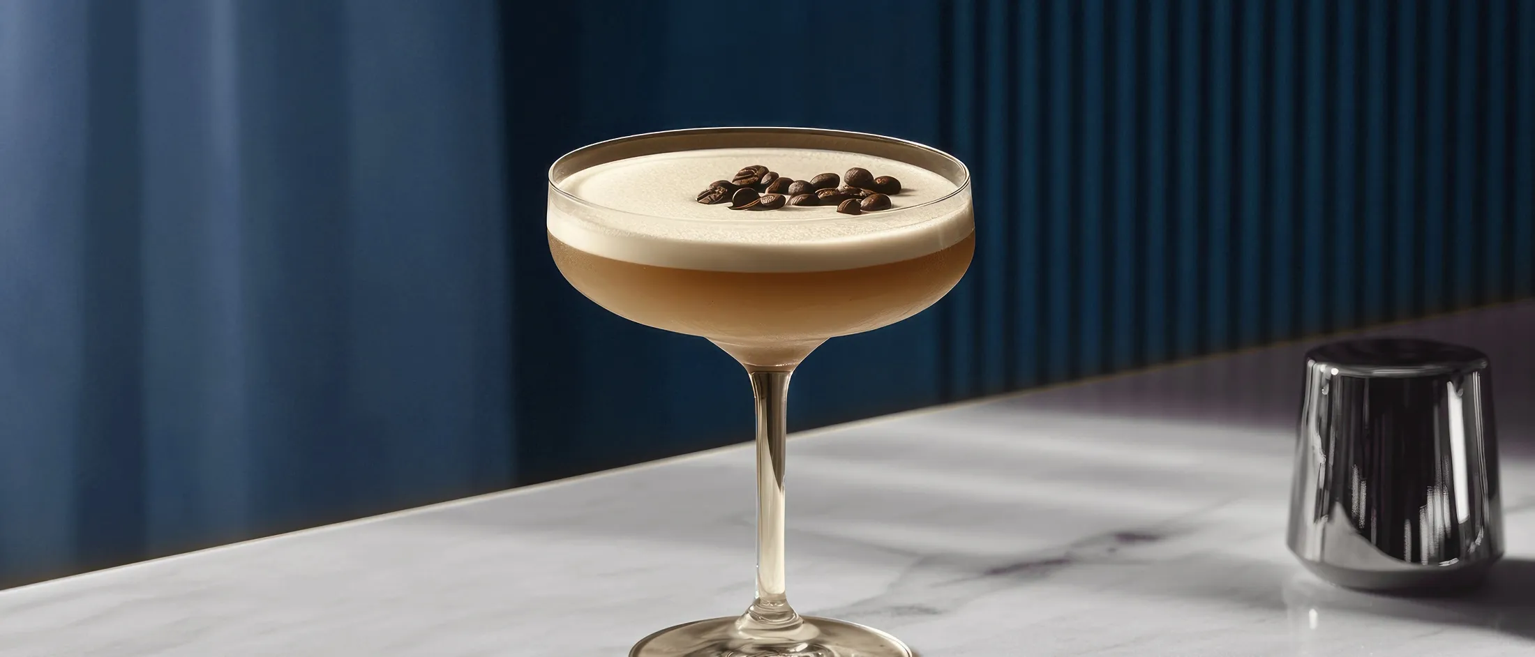 Cocktail glass with layered espresso martini topped with coffee beans on a white surface next to a small metallic container