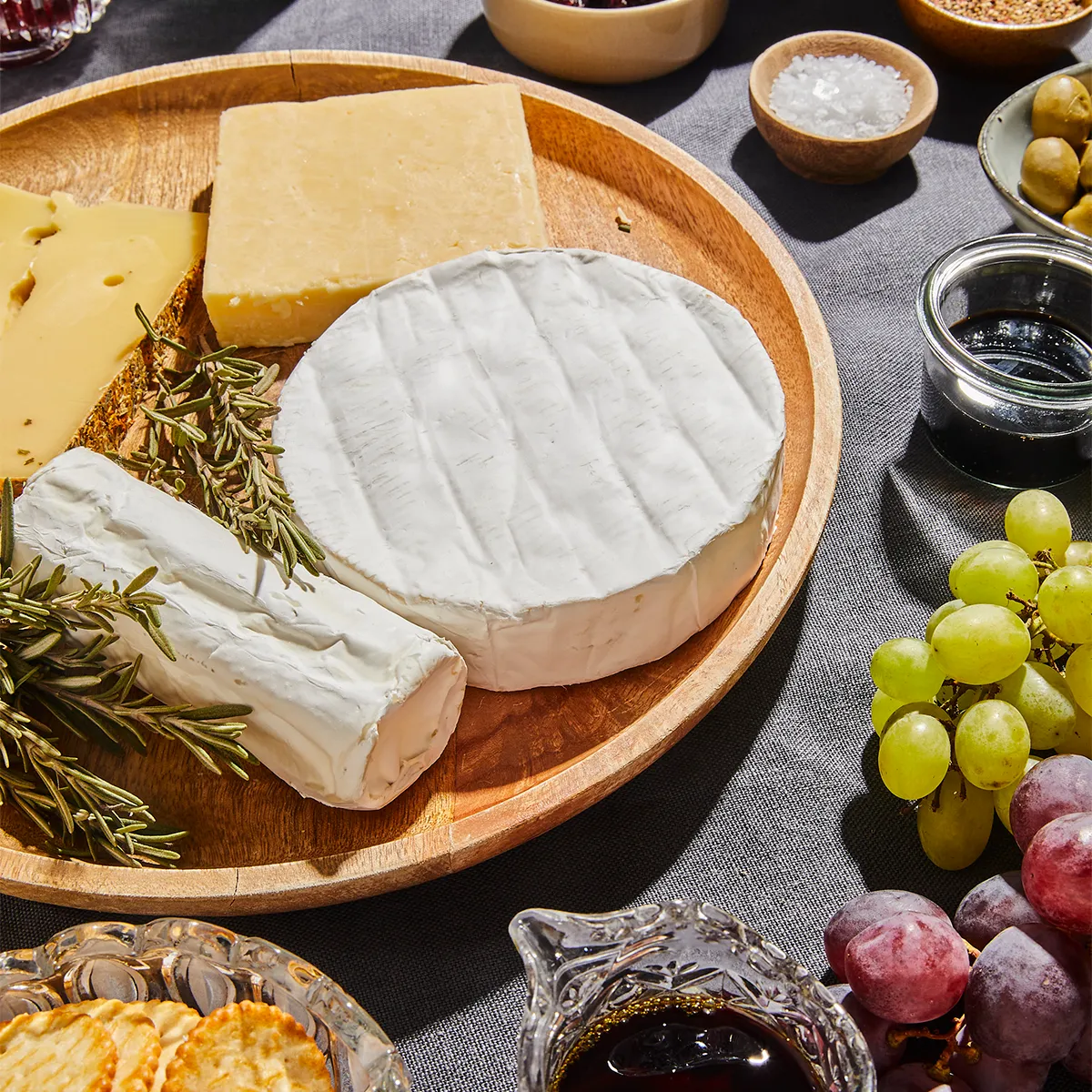 Wooden platter with assorted cheeses, grapes, crackers, olives, and rosemary on a gray tablecloth.