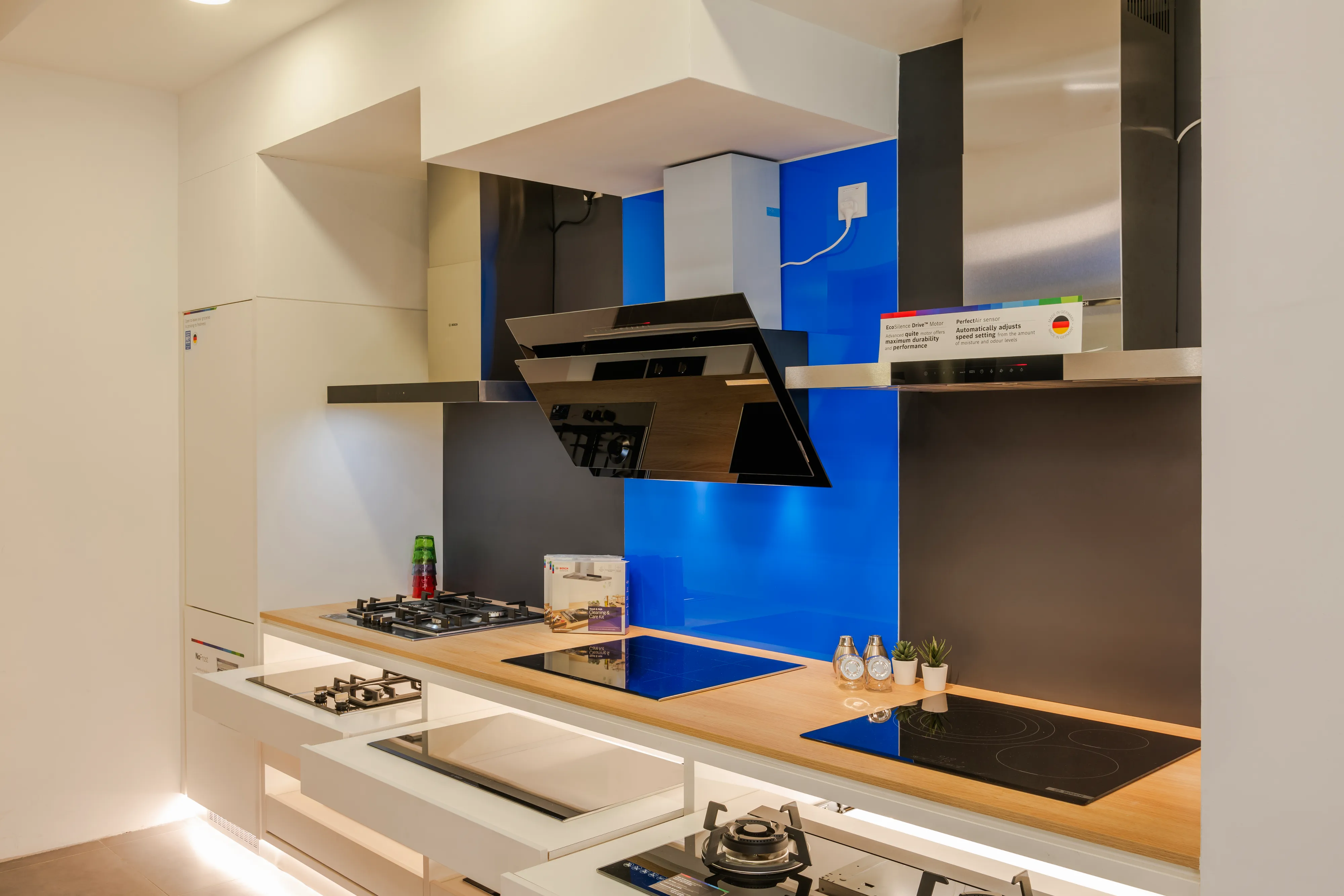 Bosch kitchen showroom display showcasing induction hobs, gas cooktops, and wall-mounted chimney cooker hoods against a blue and dark grey accent wall.