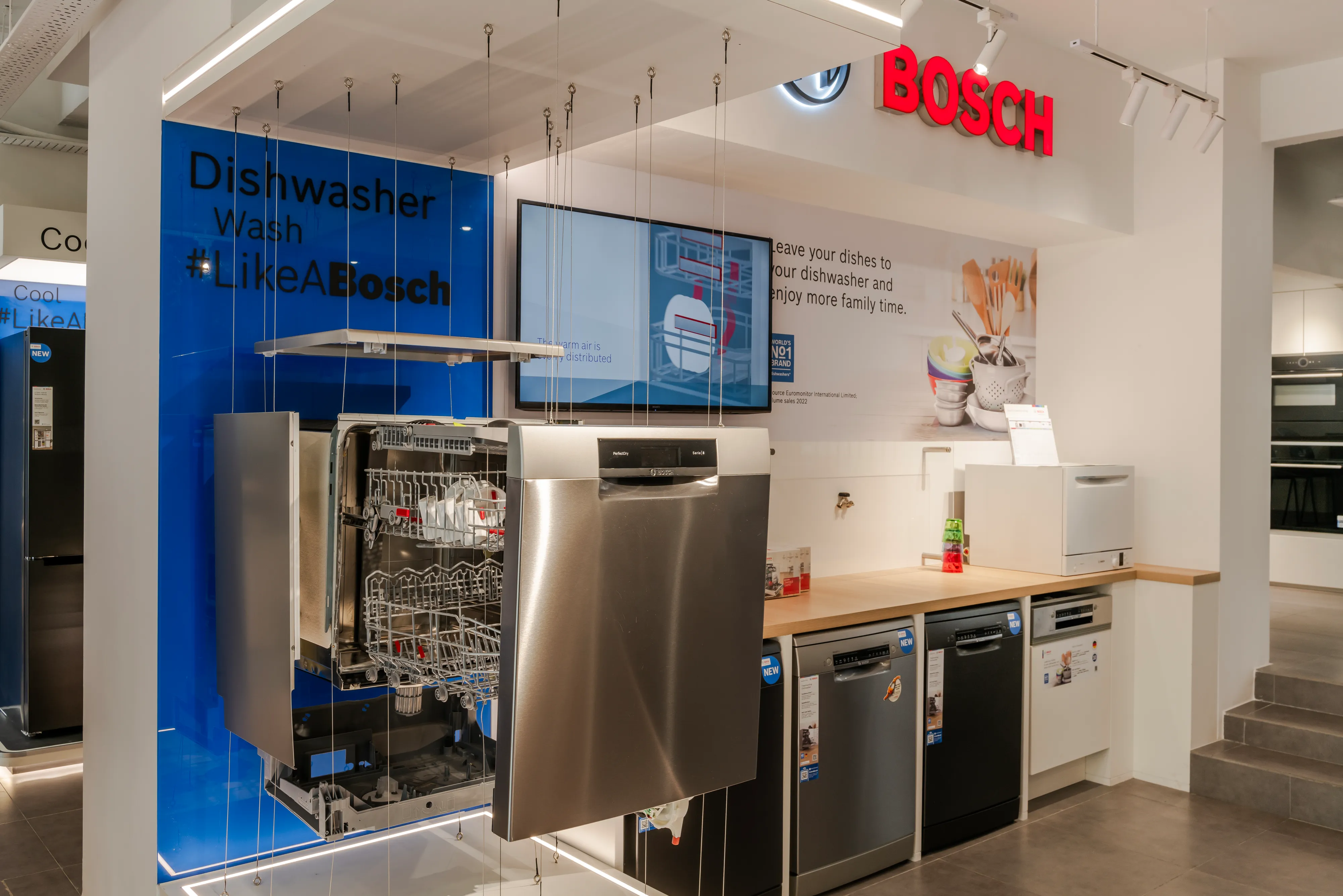 Bosch showroom dishwasher display featuring an exploded view of a stainless steel dishwasher suspended by cables alongside built-in and tabletop models under a blue "Dishwasher Wash #LikeABosch" sign.