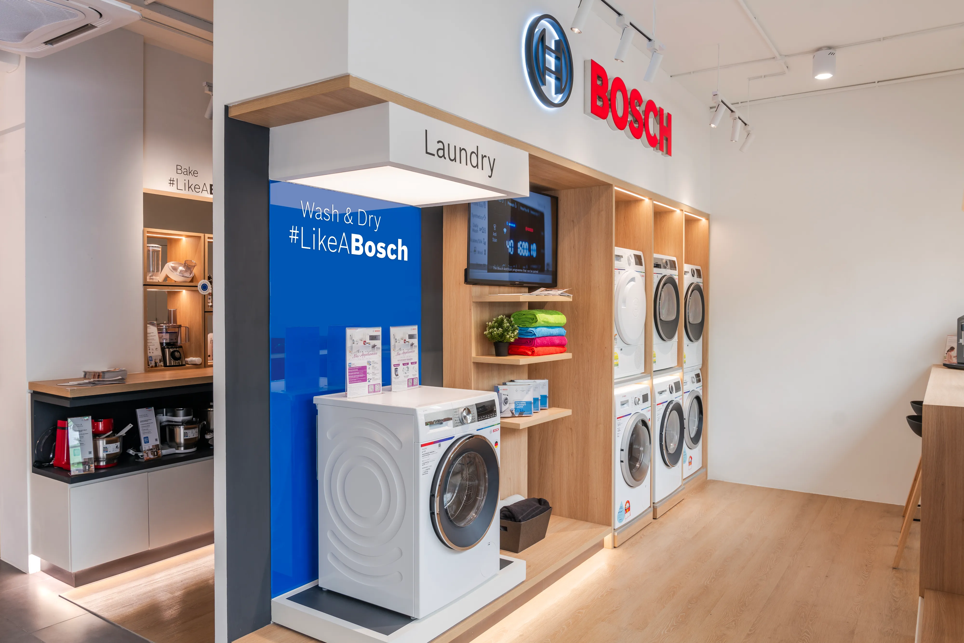 Bosch home appliance showroom section featuring front-load washing machines and dryers displayed in light wood built-in shelving, alongside a blue "Wash & Dry #LikeABosch" display.
