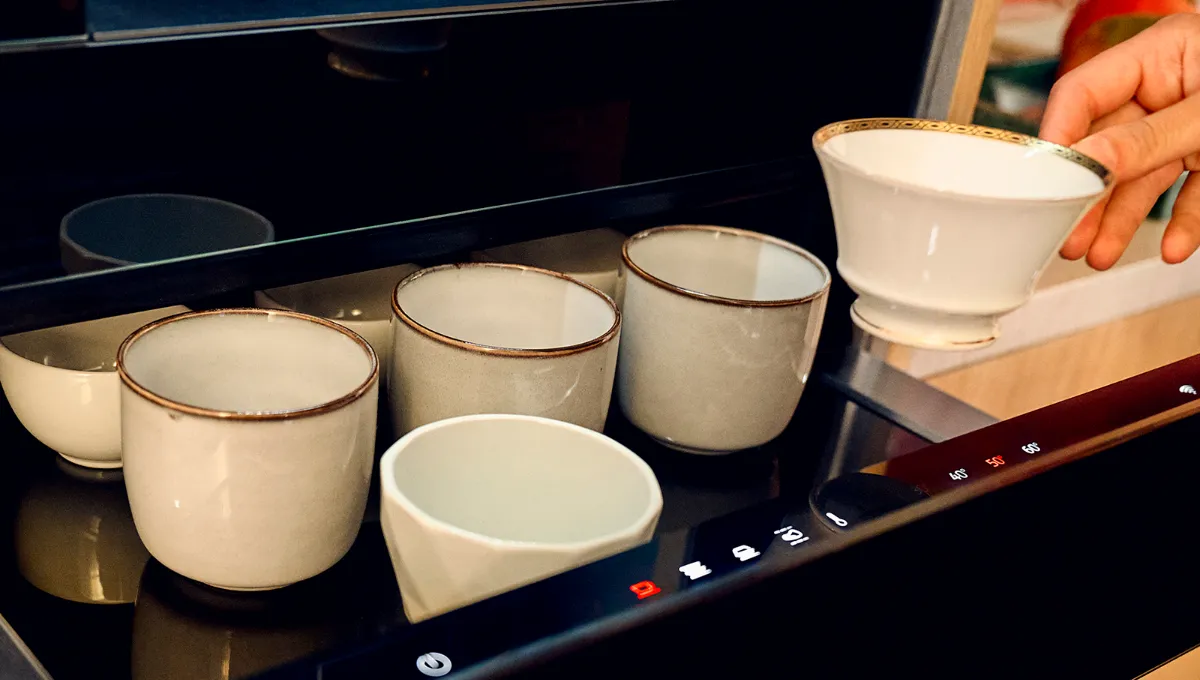 Hand placing a cup into a warming drawer with other cups and illuminated controls.