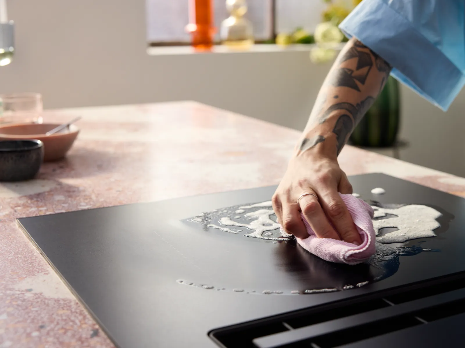 A tattooed hand cleans a black induction cooktop with a pink cloth and soapy suds.