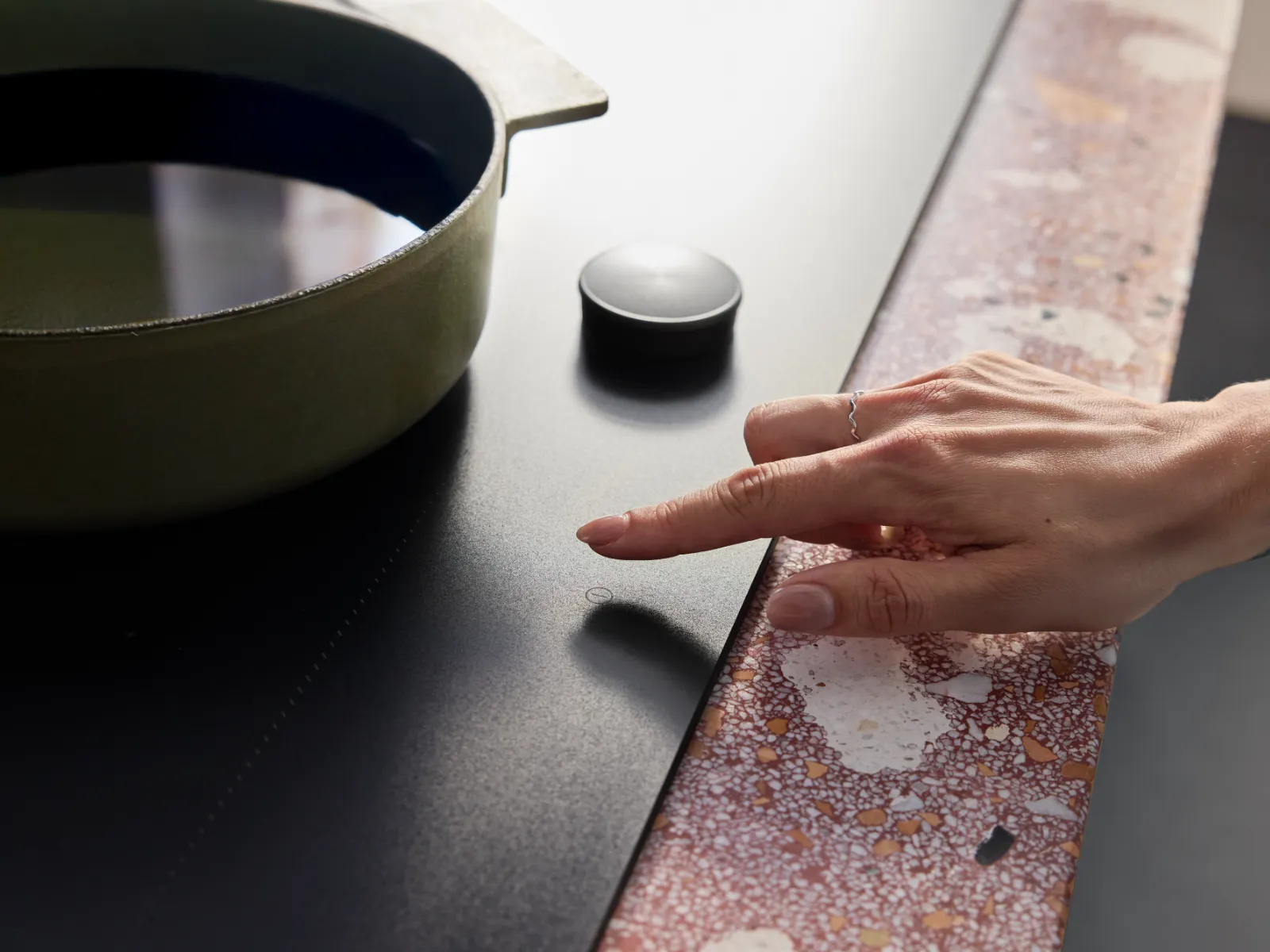 Hand adjusting the control knob on a black induction cooktop next to a green pot on a speckled countertop.