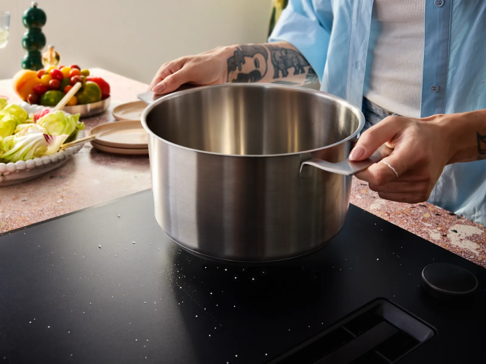 Person with tattoos placing a stainless steel pot on a black stovetop in a kitchen with fresh vegetables and glasses nearby.