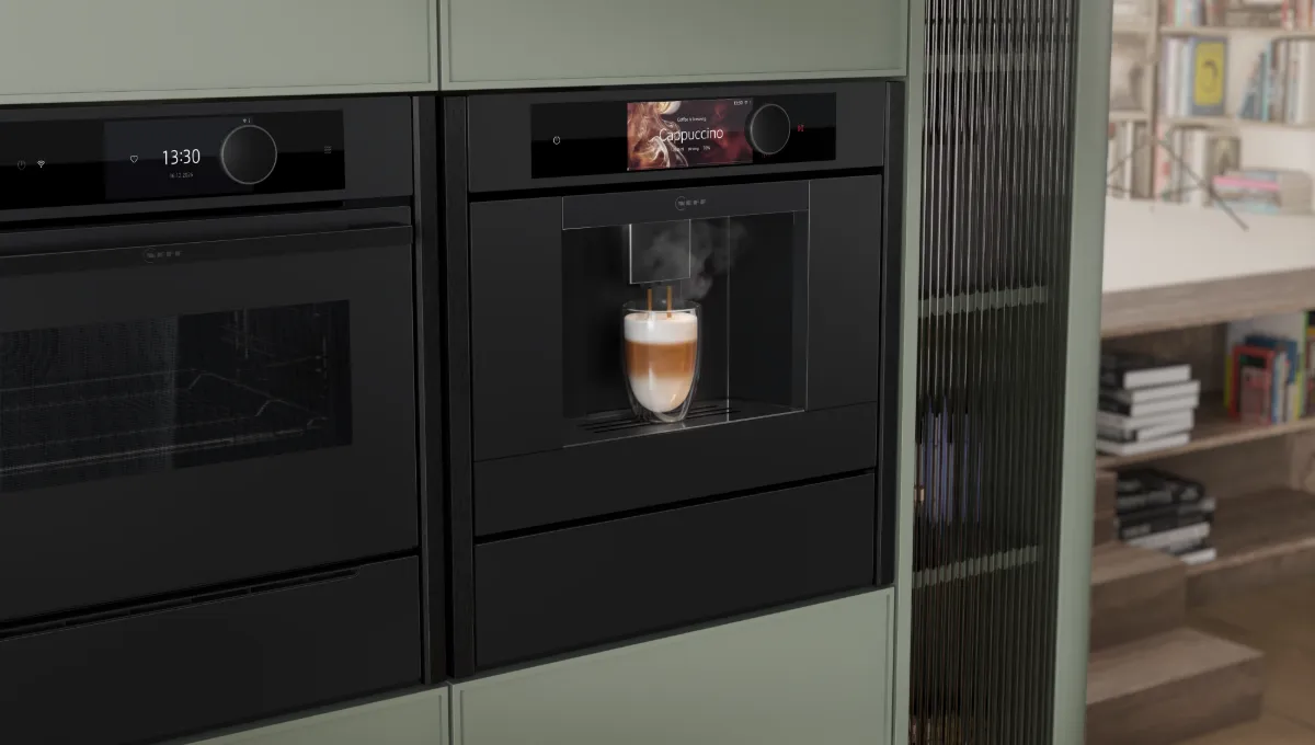 Built-in black coffee machine brewing cappuccino next to a modern oven in a green kitchen with a view of a cozy living room.