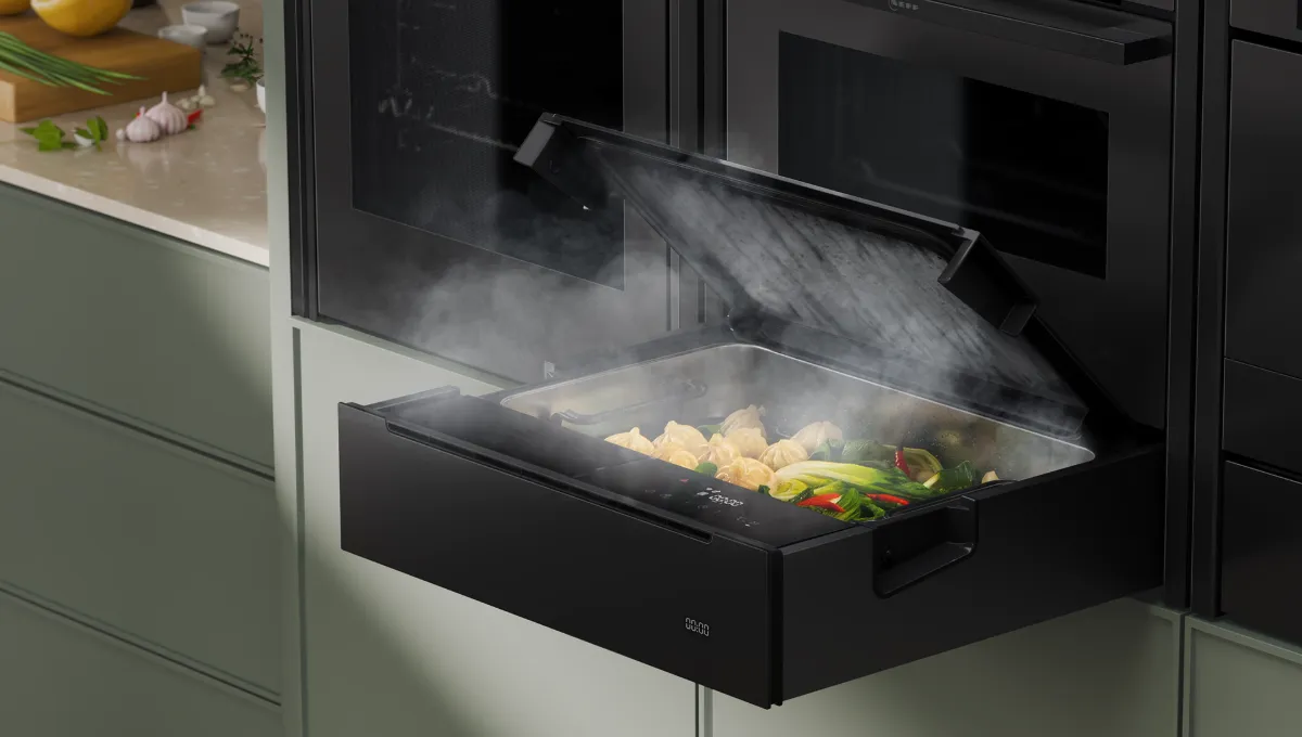 Modern kitchen with built-in steam oven drawer open, steaming cooked vegetables inside, adjacent to green cabinets and fresh ingredients on counter.