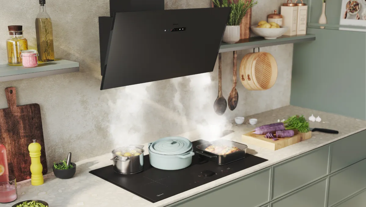 Modern kitchen counter with green cabinets, steaming pots on stovetop, hanging wooden utensils, and fresh vegetables on cutting board.
