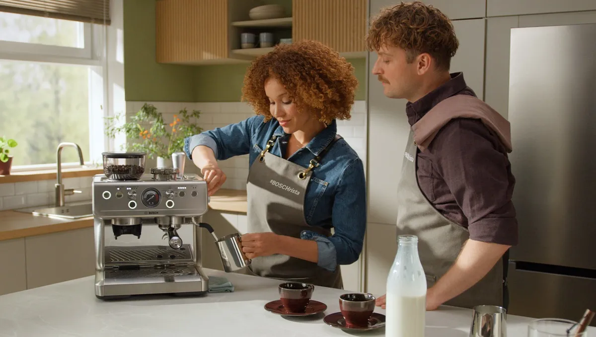 A woman in a "#BOSCHrista" apron uses a Bosch espresso machine to steam milk while a man looks on. They are in a bright, modern kitchen setting.