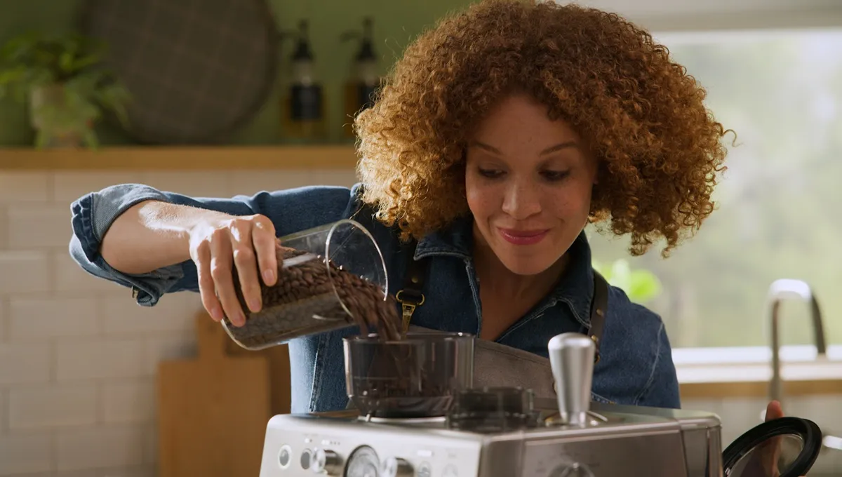 A woman with curly red hair smiles as she pours fresh coffee beans from a glass container into the grinder of a silver Bosch espresso machine in a bright kitchen.