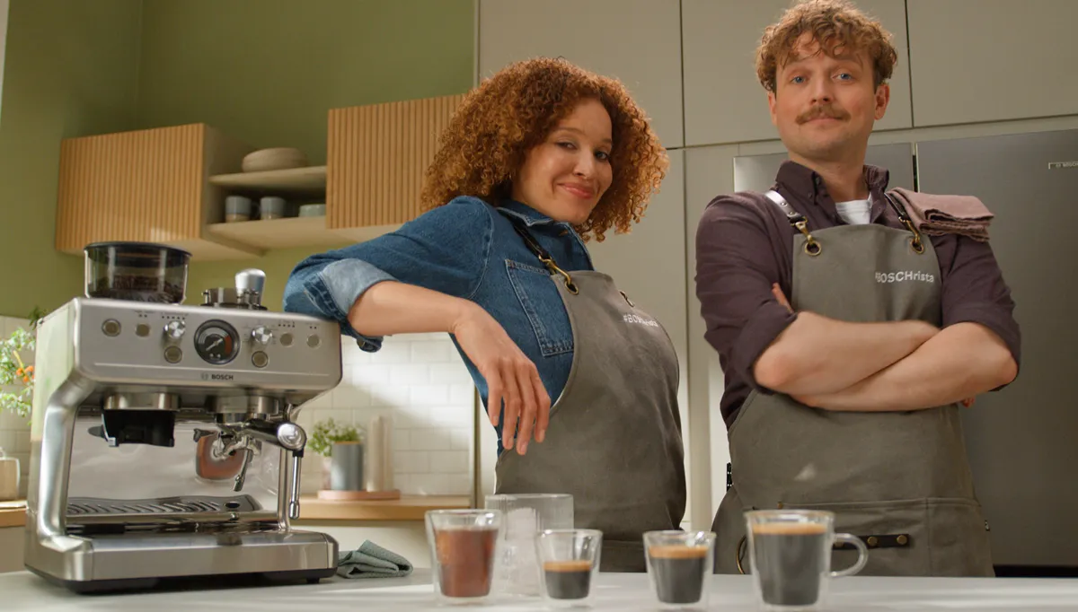 A smiling woman and a man with a mustache stand proudly behind a silver Bosch espresso machine in a modern kitchen, with four freshly made coffees ready on the counter.