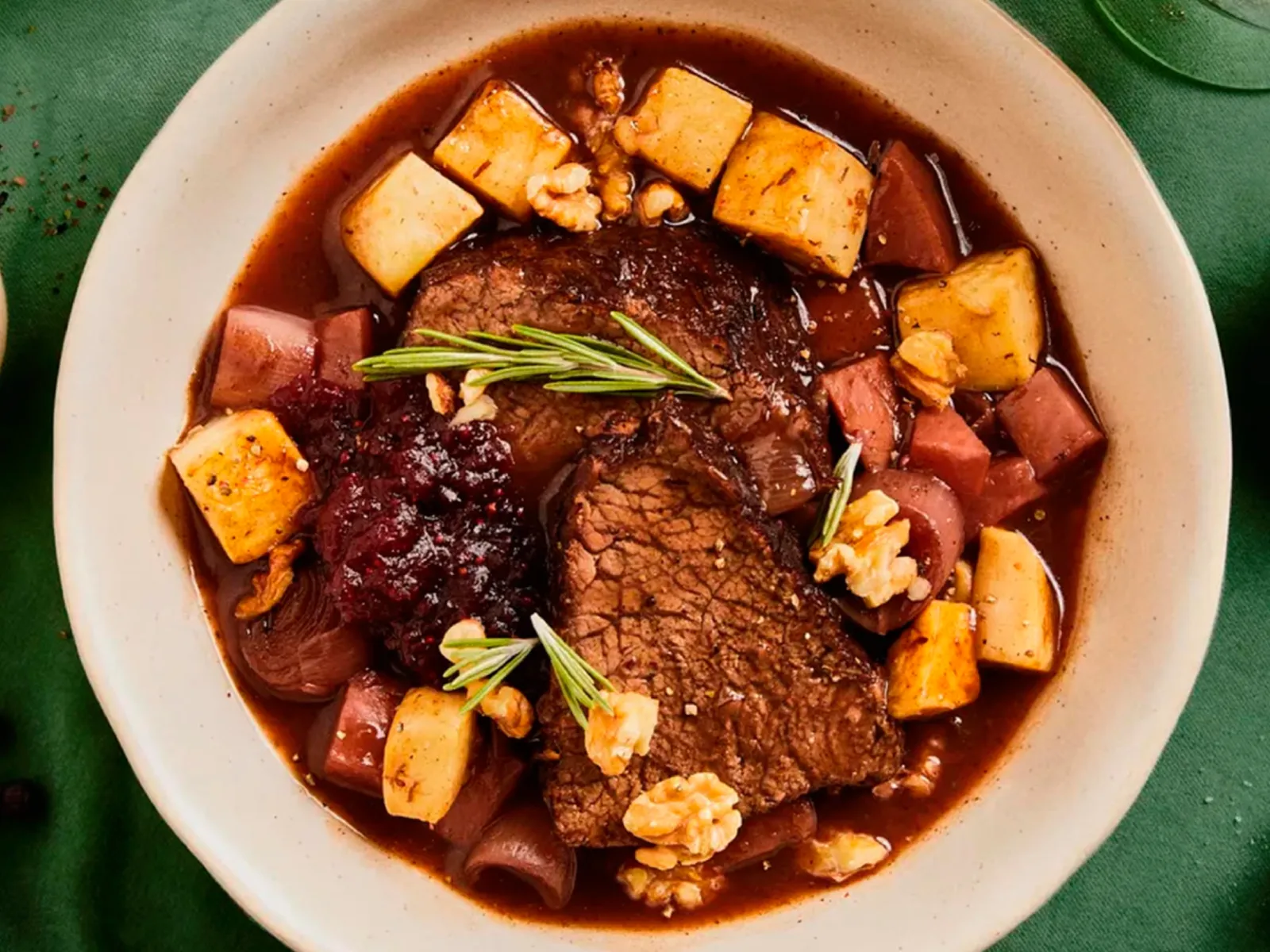 Bowl of beef stew with root vegetables, cranberries, walnuts, and rosemary on a green tablecloth with spices and herbs nearby.