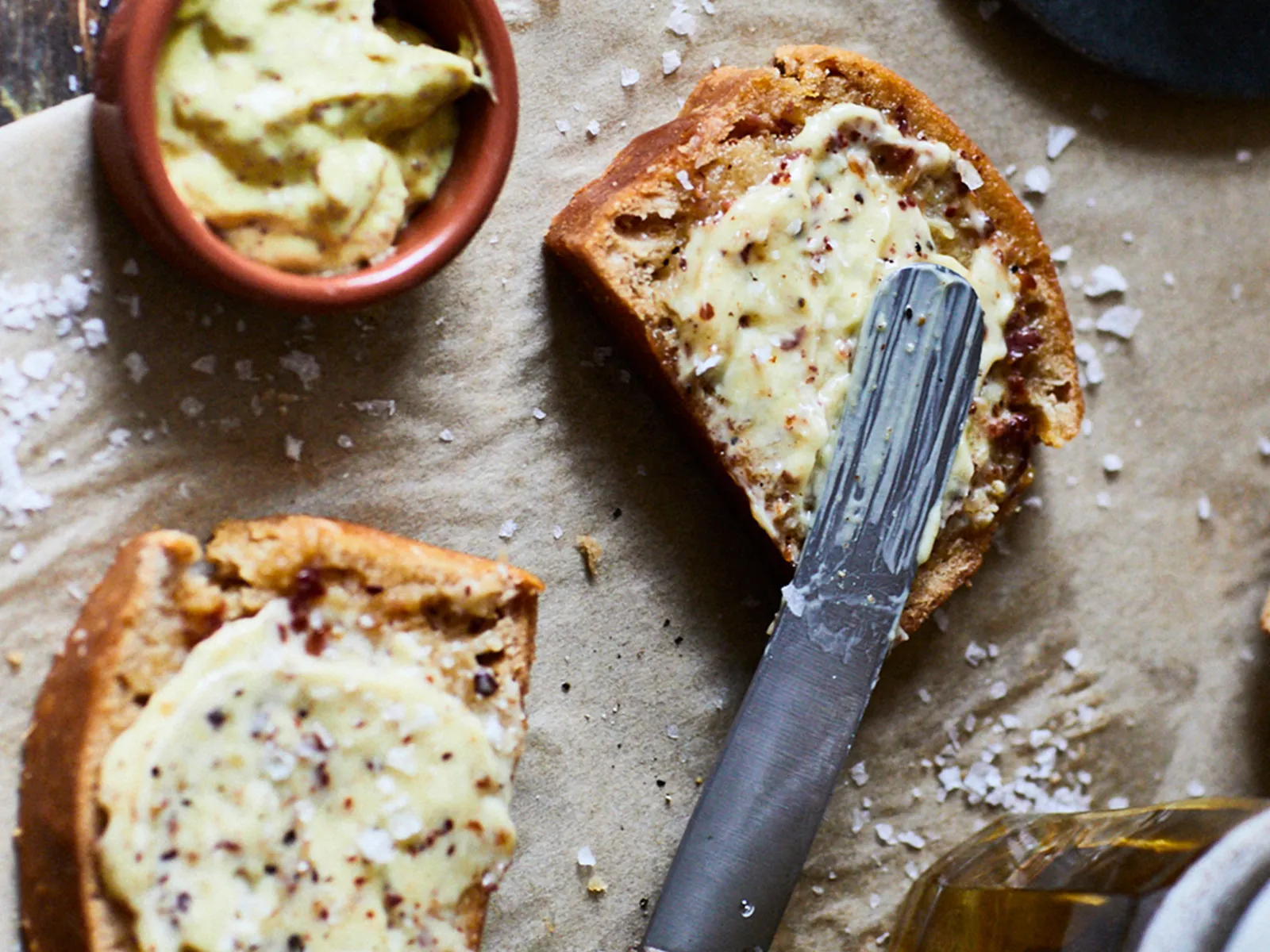 Freshly baked bread slices spread with herb butter on parchment, accompanied by a knife and a drink on a rustic wooden table.