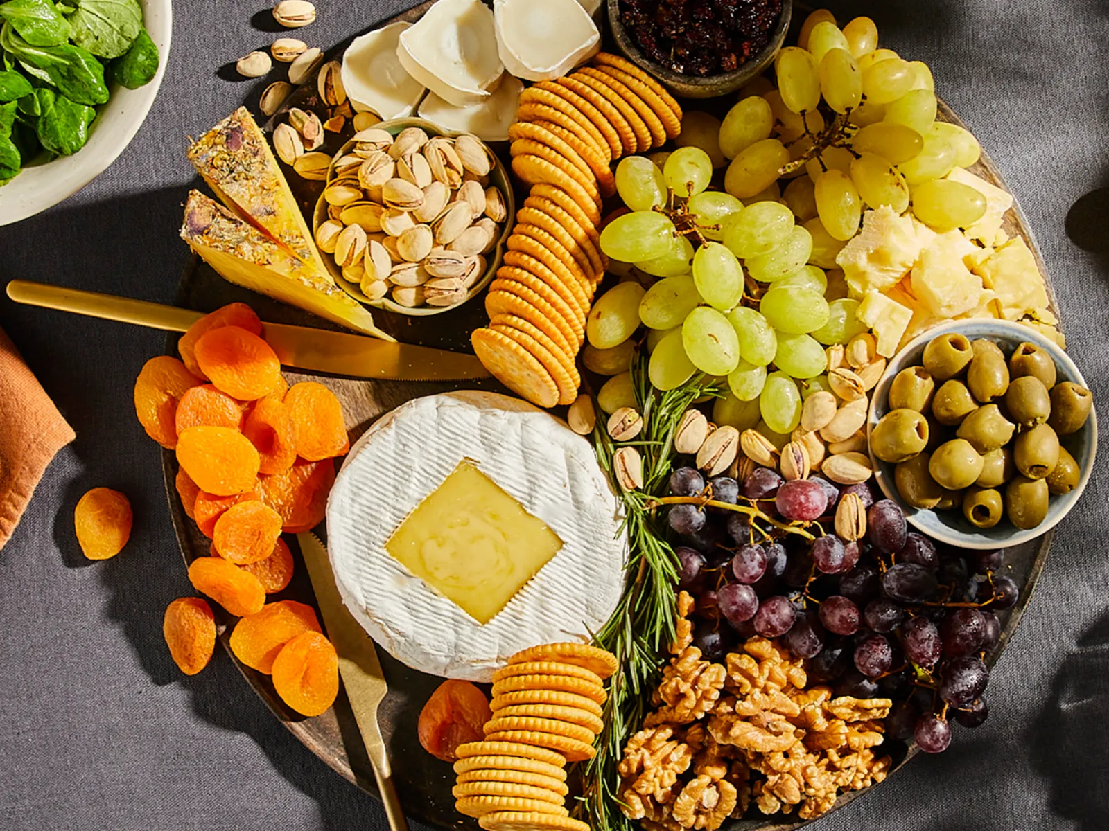 Charcuterie board with various cheeses, crackers, grapes, olives, dried apricots, pistachios, walnuts, and a bowl of spinach salad on the side.