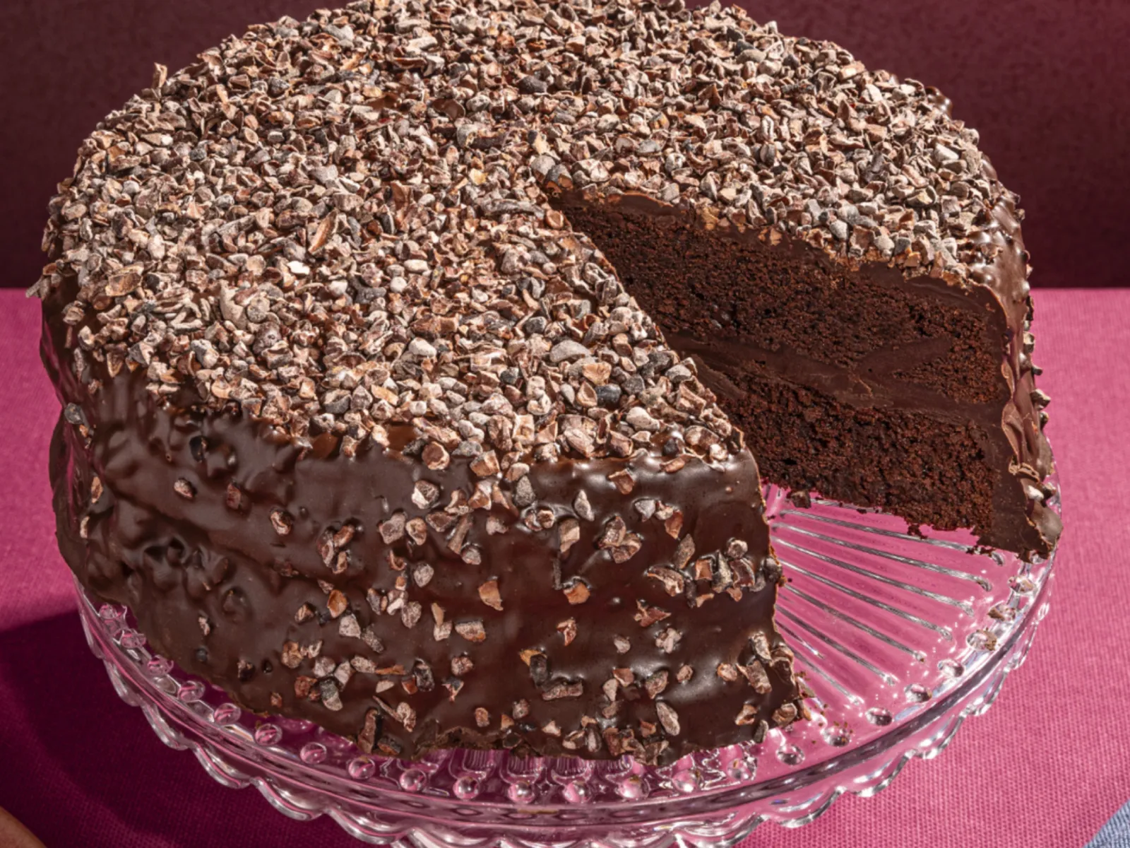 A rich chocolate cake, covered in chocolate chips, sits on a glass cake stand with a slice removed.