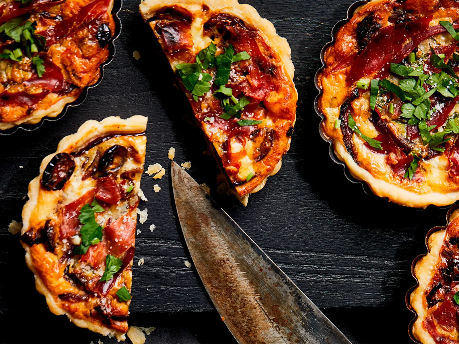 Five savory tarts with herbs and pepperoni displayed on a dark slate surface with a knife.