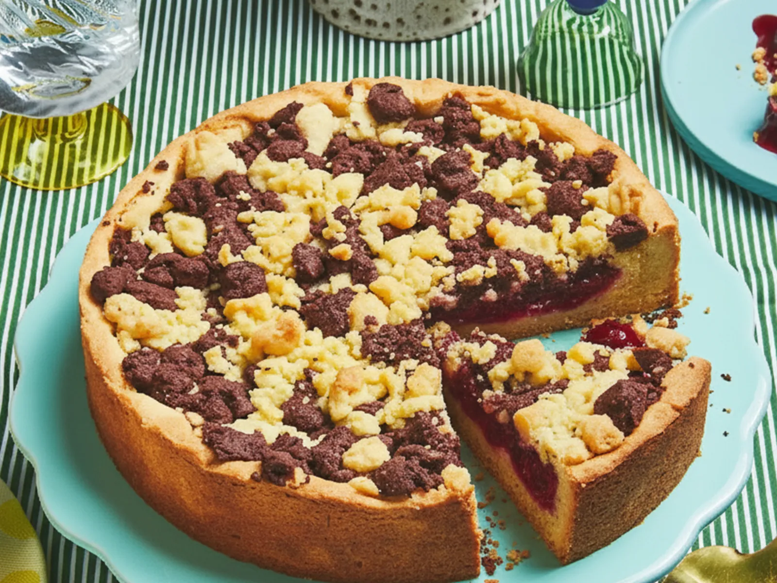 A sour cherry streusel cake sits on top of a light blue cake stand on a green tablecloth and is surrounded by other tableware.