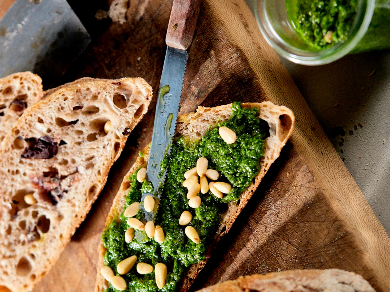 Sliced bread on a wooden board, topped with pesto and pine nuts, knife resting beside.