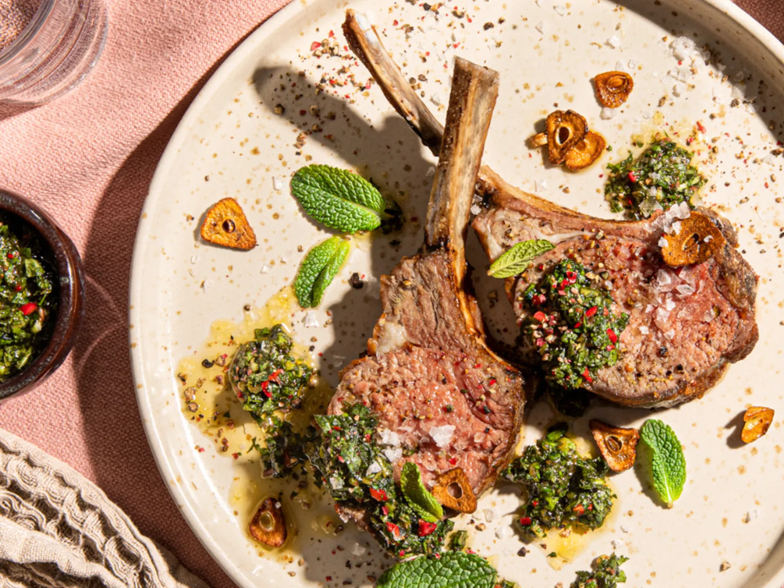 A plate with two grilled lamb chops topped with herbs, garlic chips, and a vibrant green sauce, surrounded by garnishes and a glass of water.