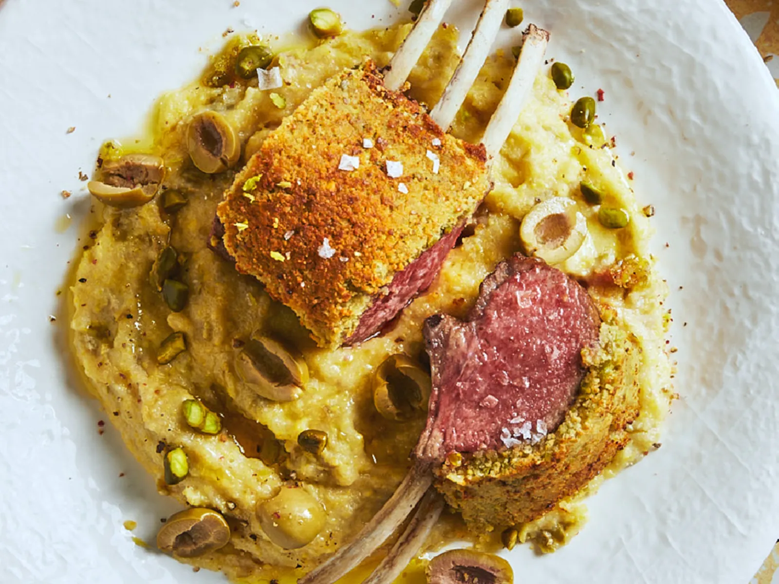 A pistachio crusted rack of lamb sits on polenta on a white plate placed on yellow ceramic tiles and is surrounded by other tableware.