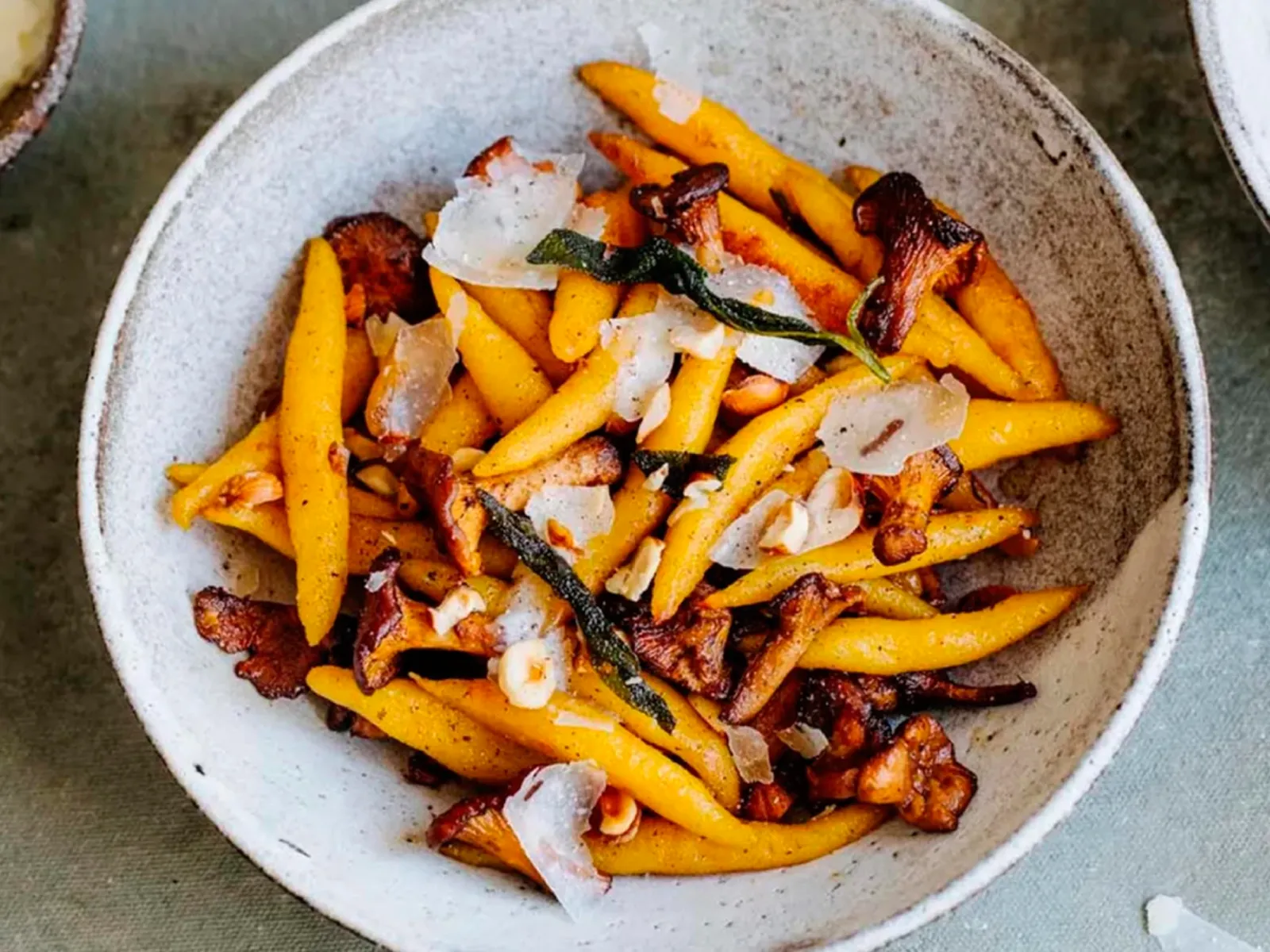 Plate of golden pasta with sautéed chanterelle mushrooms, sage leaves, shaved cheese, and crushed nuts on a gray surface.