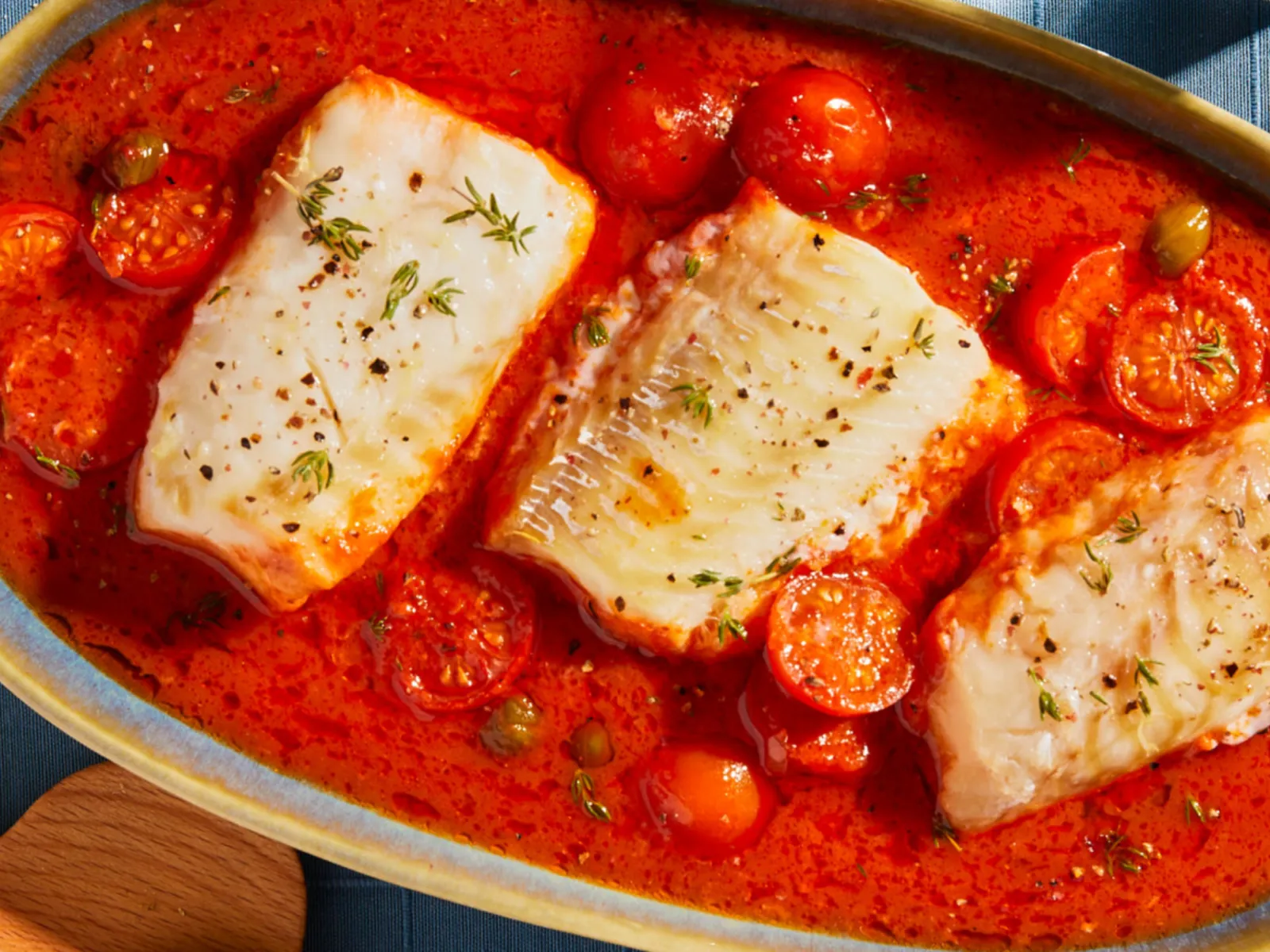 Baked white fish fillets in a tomato-based sauce with cherry tomatoes and herbs, served in an oval ceramic dish.