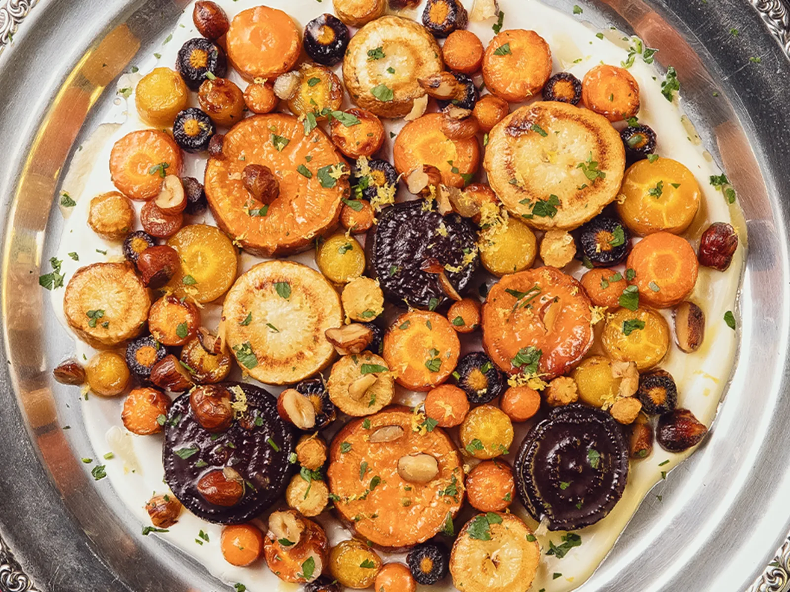 Colorful roasted root vegetables cut into rounds, arranged on top of a yogurt base on a serving platter and topped with brown butter and hazelnuts.