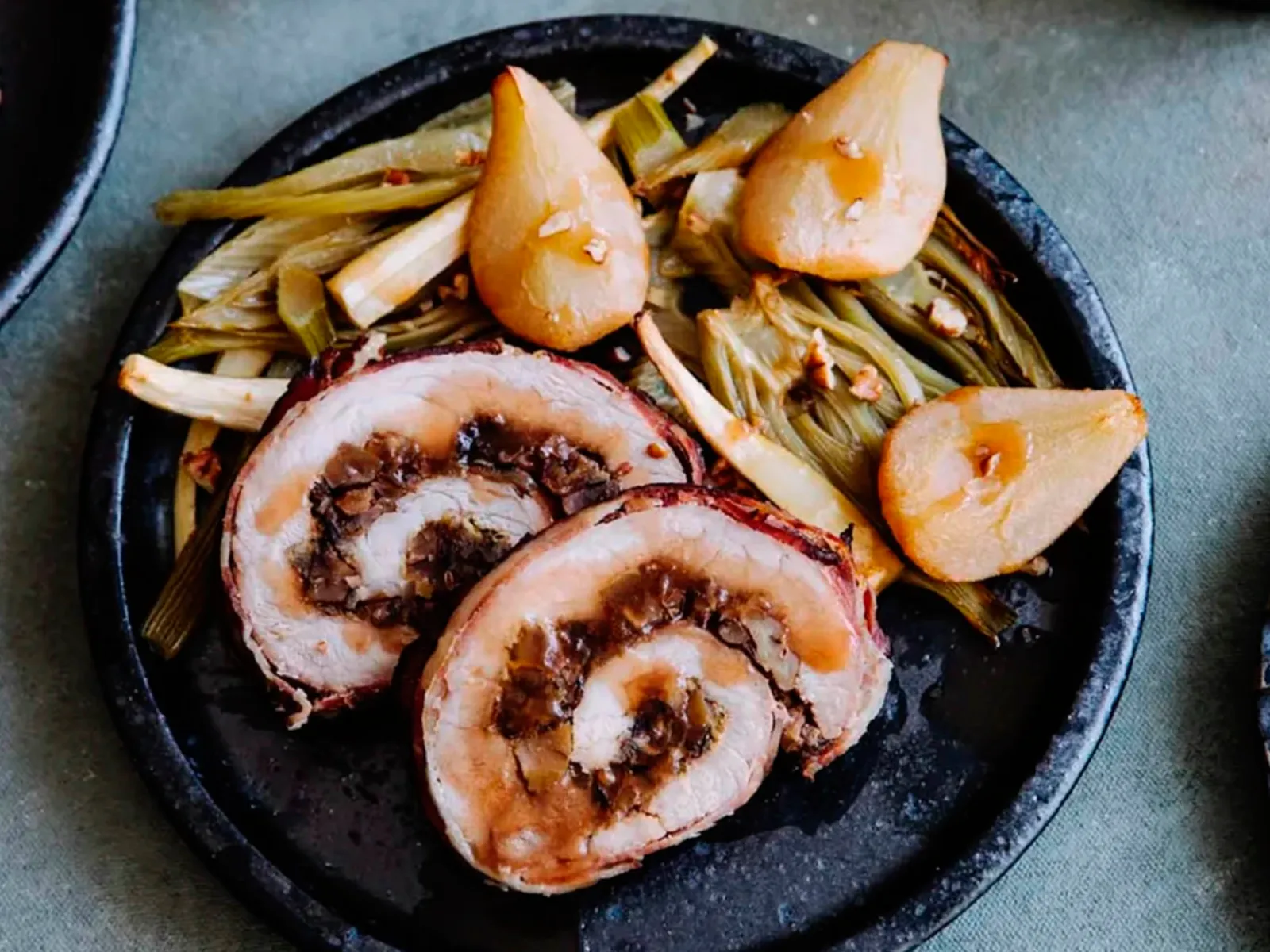 Sliced rolled pork roast with stuffing served with braised leeks and pears on a dark plate.