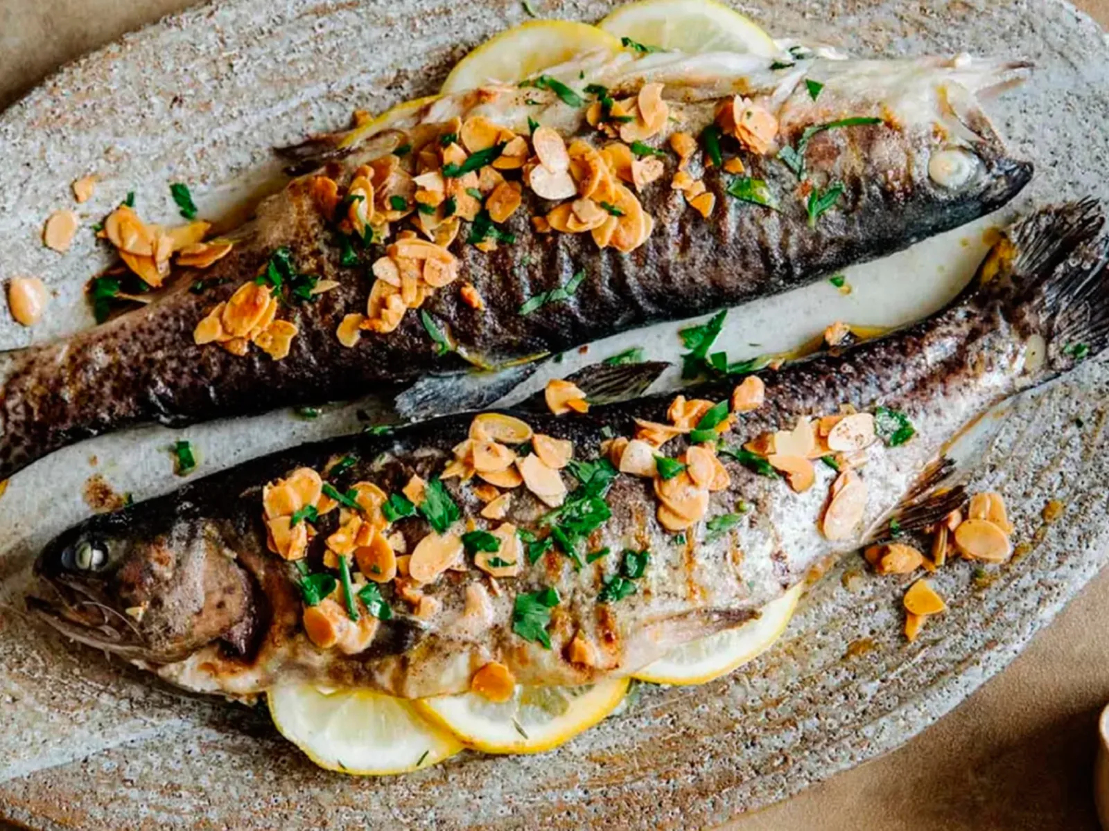 Two whole grilled fish garnished with sliced almonds and chopped herbs, served on lemon slices on a rustic oval platter.