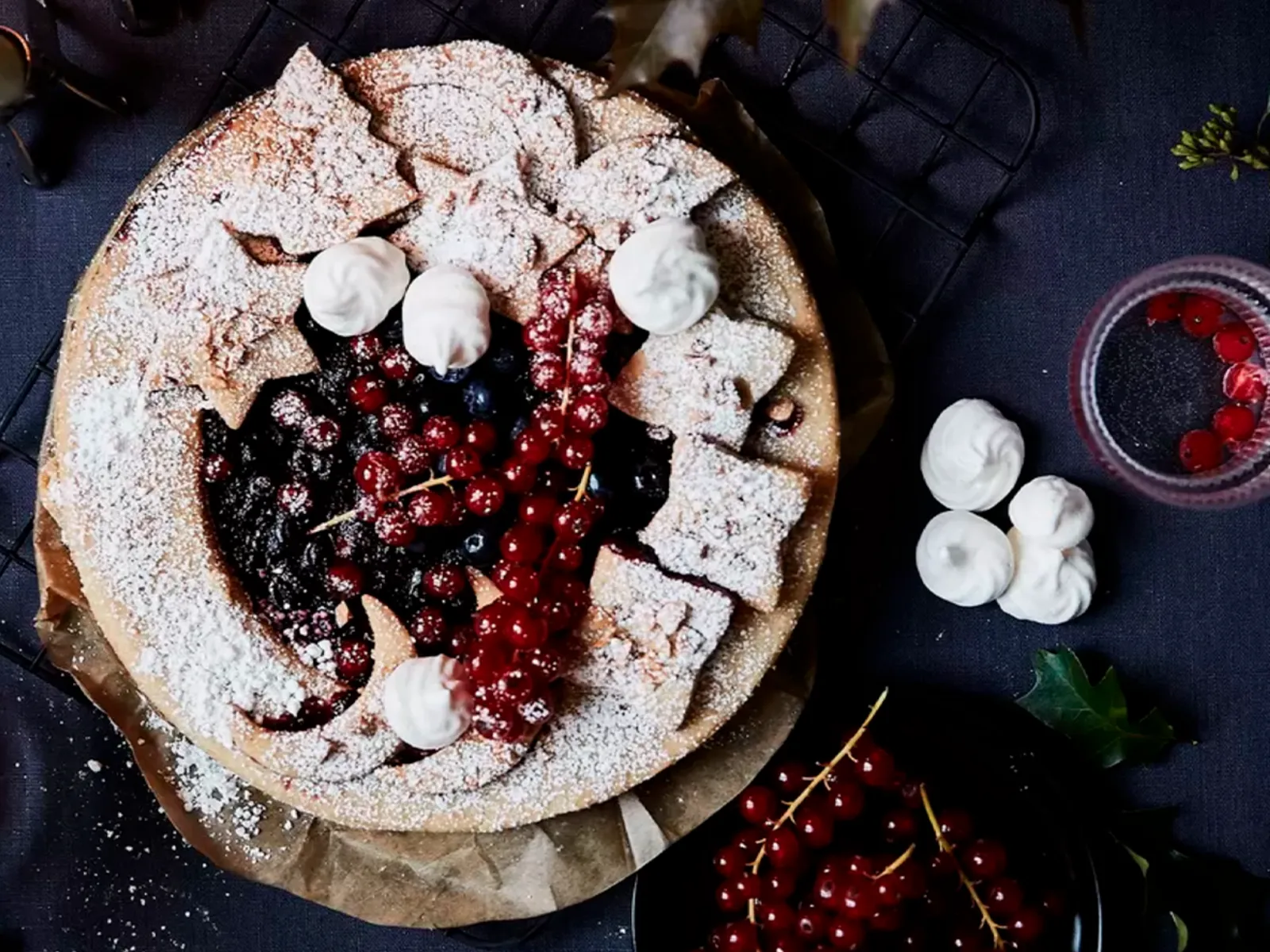 Top-down view of a berry tart dusted with powdered sugar, surrounded by plates with meringues, red currants in glasses, and a lit candle.
