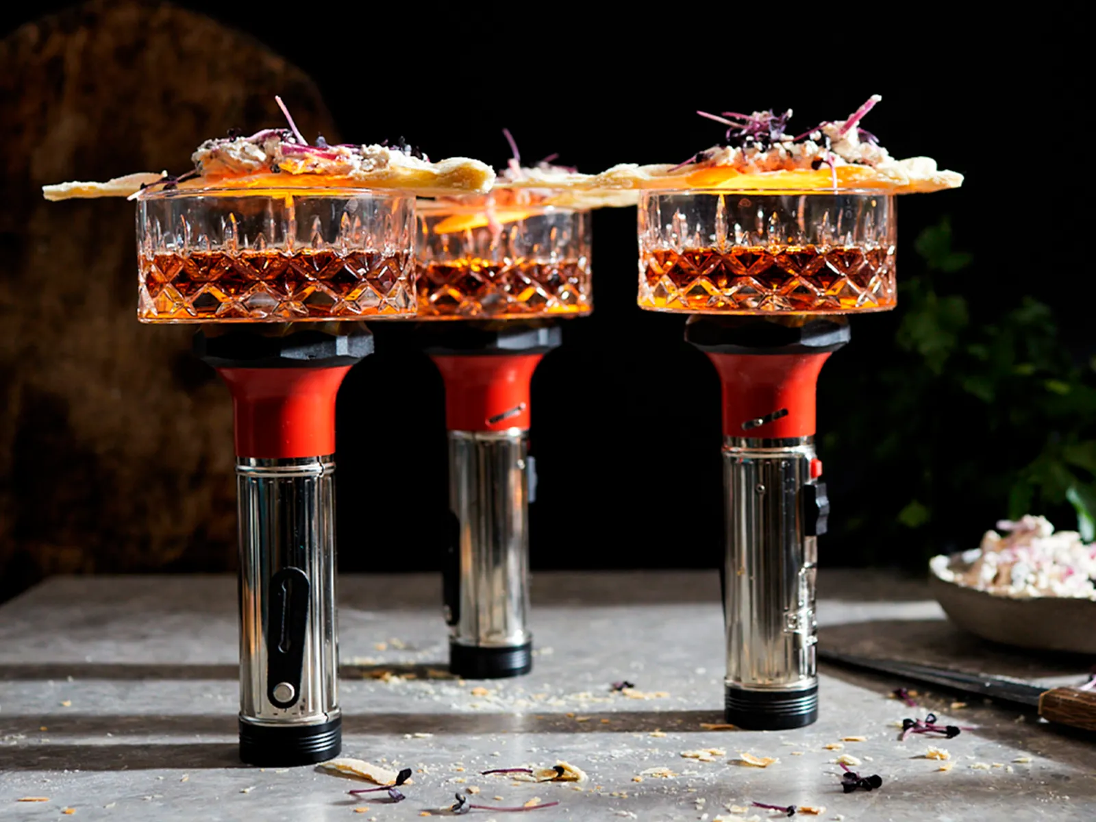 Cocktail glasses filled with amber liquid, garnished with citrus slices, on a dark background with various bar tools.