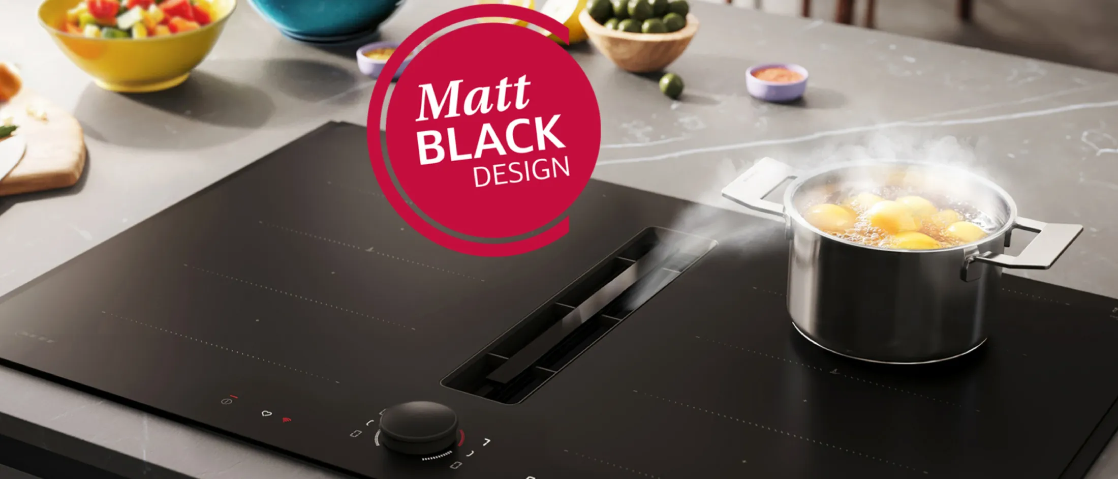 Black matte induction cooktop with a central downdraft vent and a round control knob; a steaming stainless-steel pot of boiling eggs sits on the right burner. Bowls of salad and olives sit on the countertop behind, and a red circular badge near the top reads "Matt BLACK DESIGN."