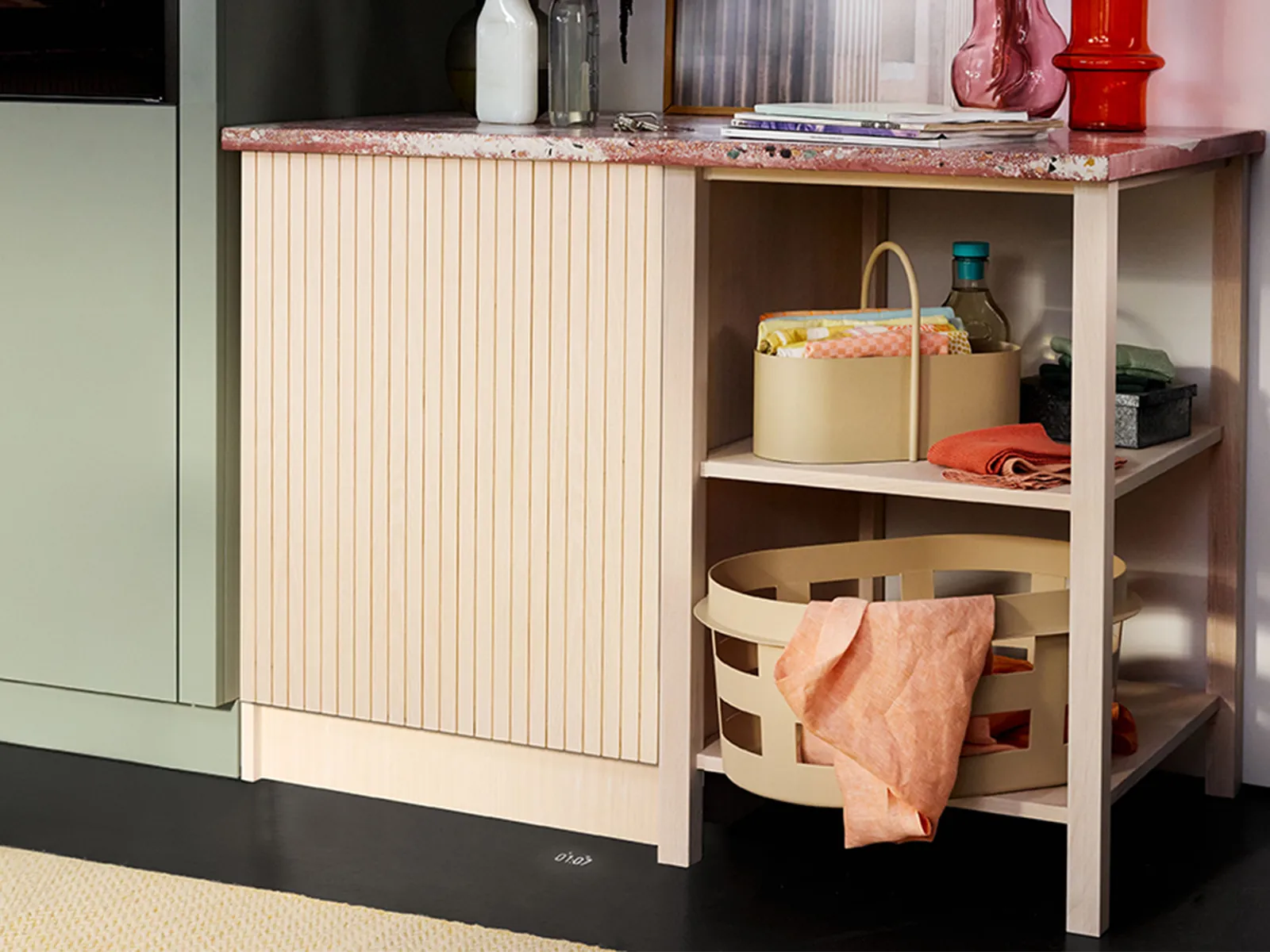 A stylish kitchen corner featuring a textured beige cabinet, marble countertop, and organized baskets with vibrant fabrics and kitchen items.