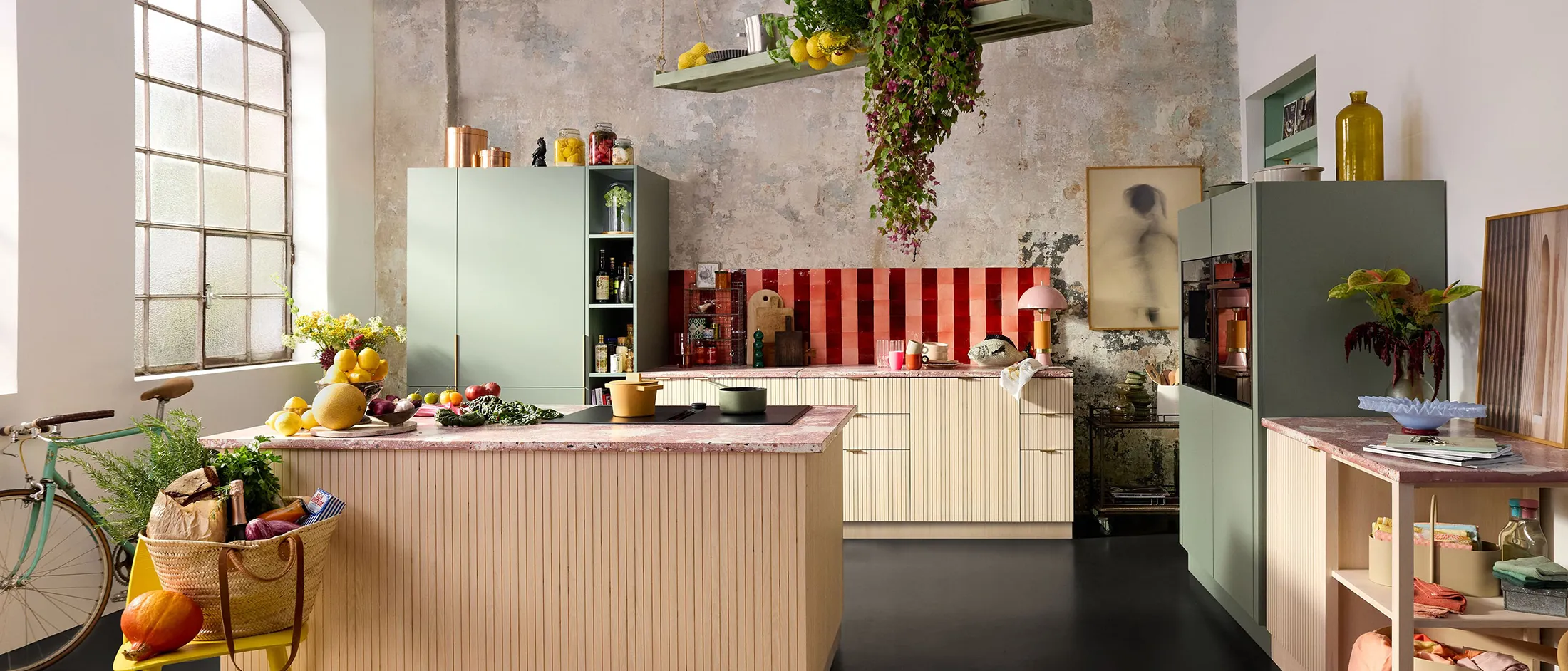 Bright, modern kitchen featuring green cabinetry, a vibrant red backsplash, abundant produce, and a bike leaning against the wall.