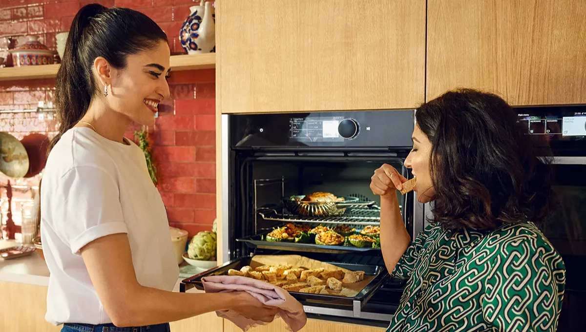Two smiling women in a modern kitchen. One samples food from an oven tray, while the other holds a tray of freshly baked items.