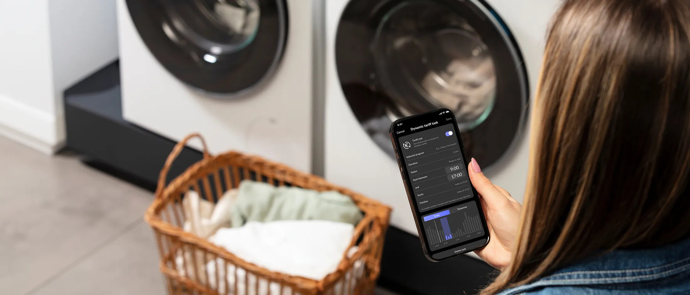 Femme utilisant le smartphone tout en faisant la lessive, machine à laver et panier en arrière-plan.