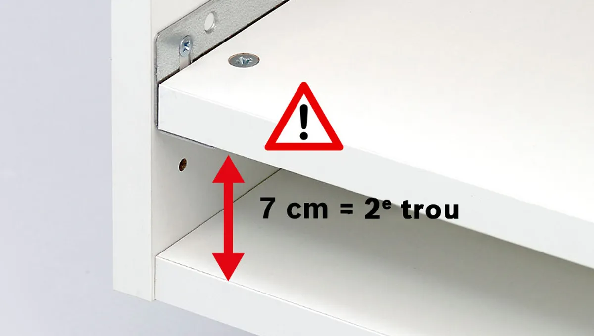 Nærbilde av hjørnet på en hvit hylle med en metallbrakett, som viser et varselskilt og en avstand på 7 cm mellom to borede hull.
