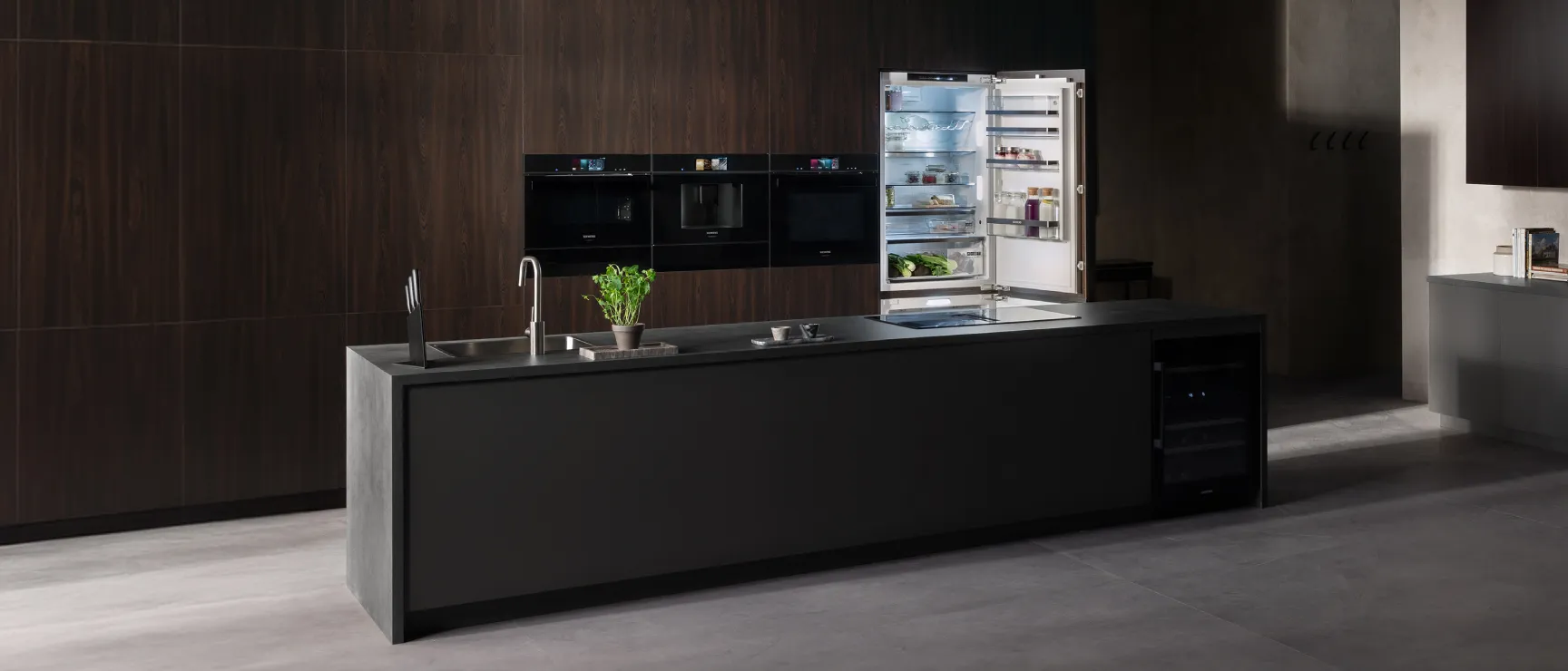 A wide shot of a luxurious, modern kitchen with dark wood-paneled walls and a large, dark grey central island. The background features a bank of black Siemens built-in ovens and a tall, integrated refrigerator with its door open.