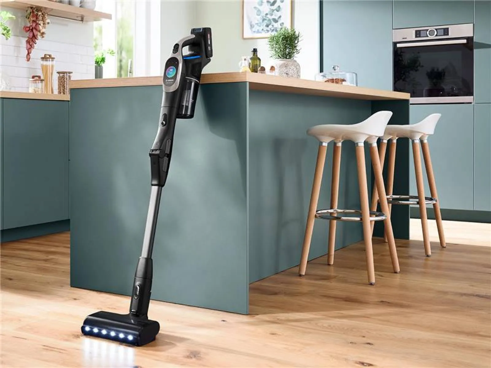 Black Bosch Unlimited 7 cordless stick vacuum leaning against a teal kitchen island, LED-lit motorized floor head on a hardwood floor beside two white bar stools.