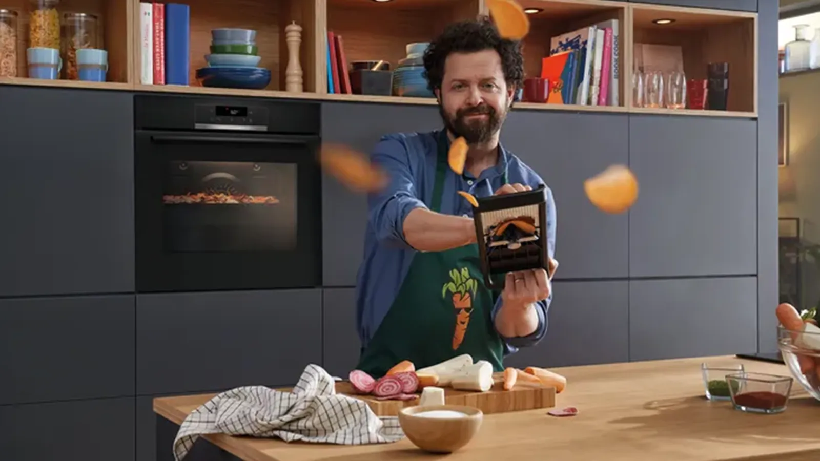 Ein lächelnder Koch mit Schürze wirft in einer modernen dunklen Küche mit Holz-Arbeitsplatte und Bosch Einbaubackofen Gemüsechips in die Luft.