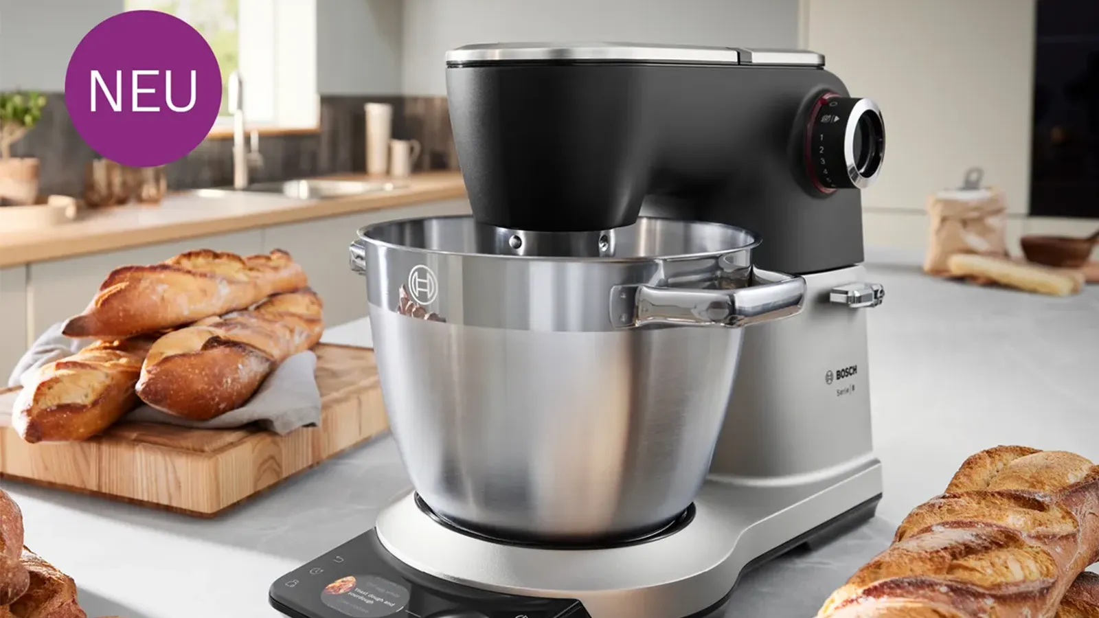 Eine moderne, mattschwarze und silberne Bosch Küchenmaschine mit großer Edelstahlschüssel auf der Arbeitsplatte, umgeben von frisch gebackenem Brot, inklusive lila „NEU“-Siegel.