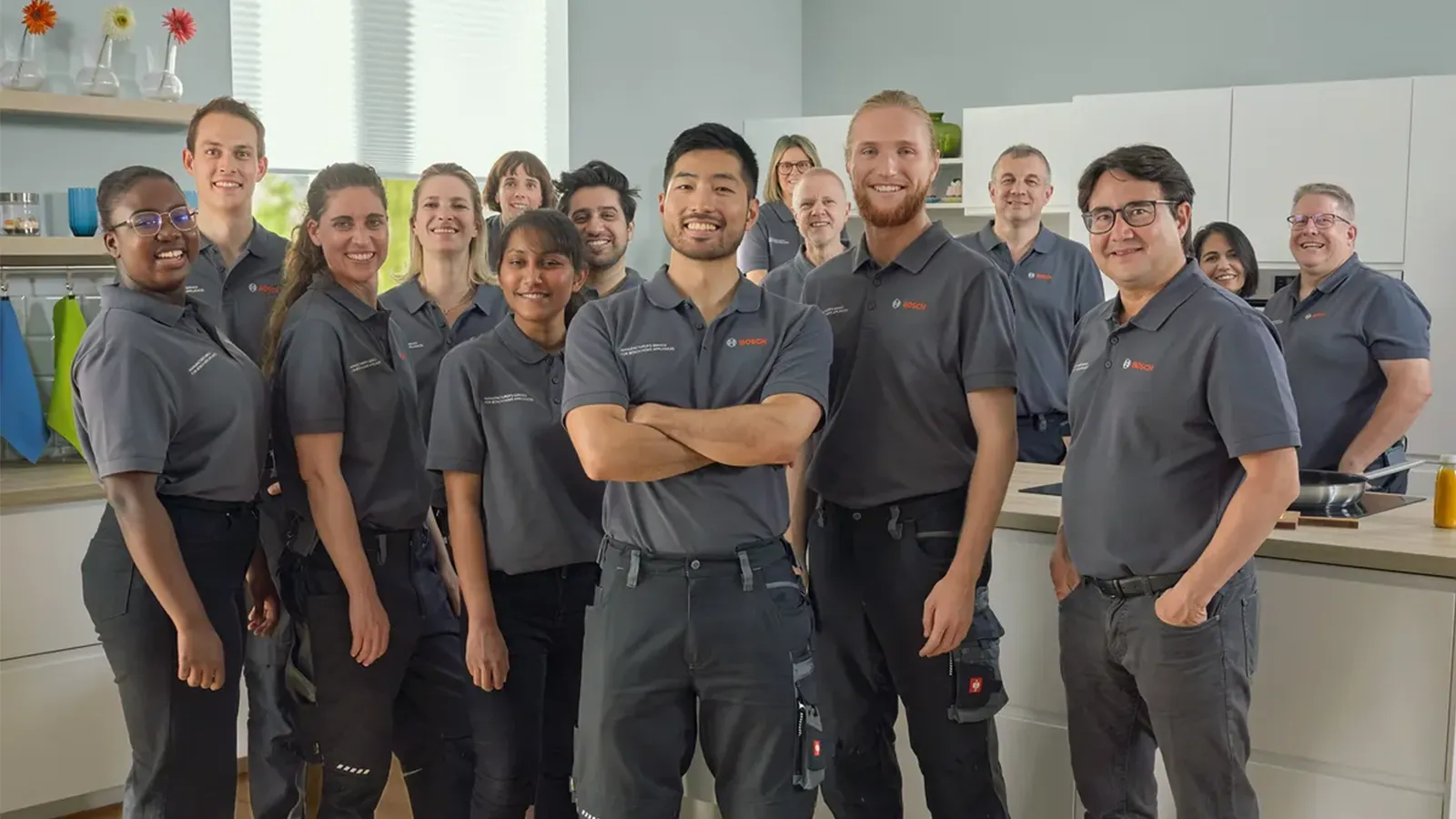 Ein großes, vielfältiges Team von Bosch-Mitarbeitern in grauer Arbeitskleidung posiert lächelnd in einer modernen Studio-Küche.