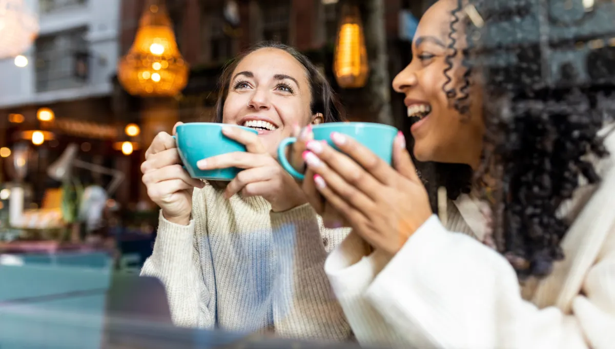 Two women smiling and enjoying hot drinks together in a cozy, warmly lit café setting.