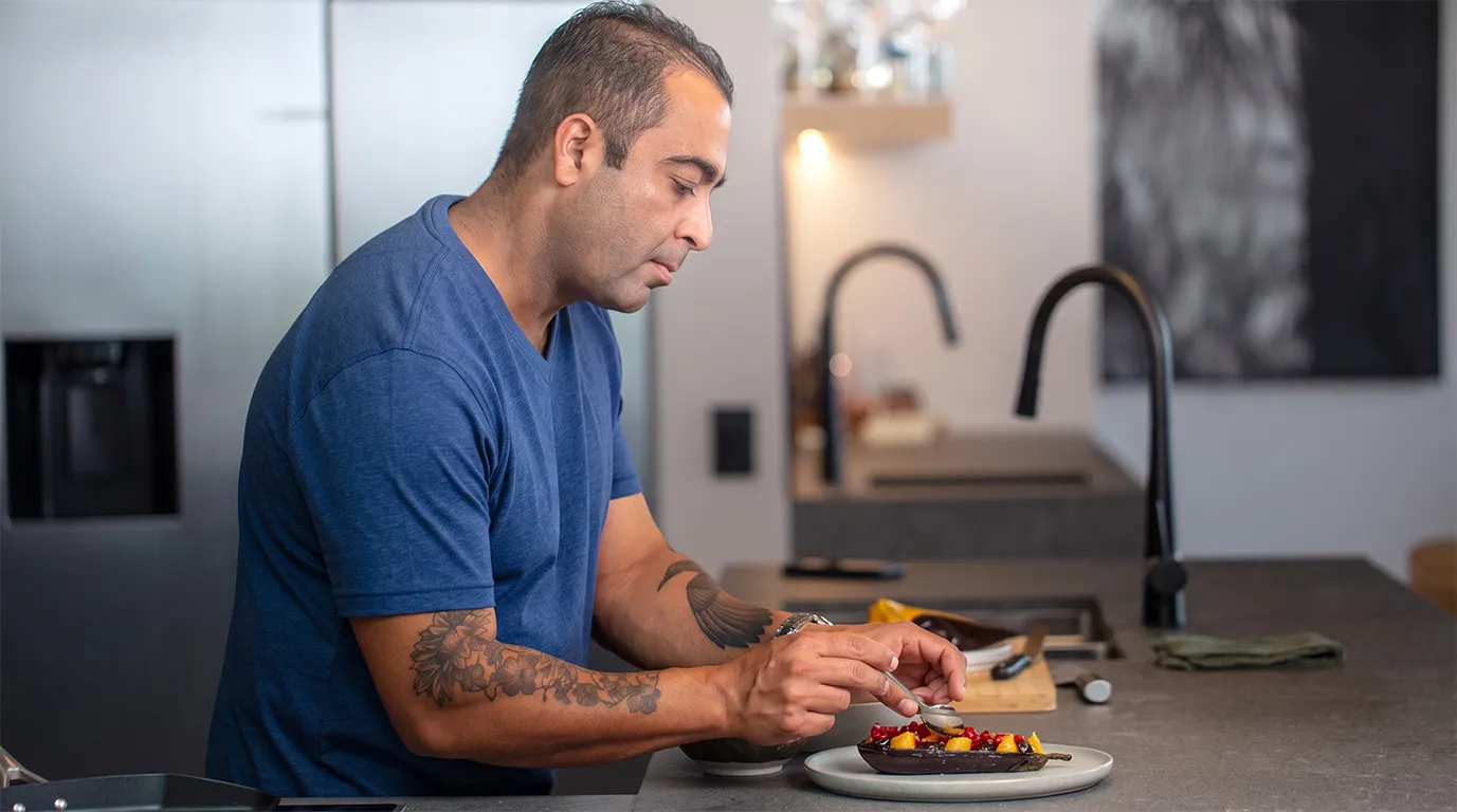 A man with dark hair and tattooed forearms, wearing a blue t-shirt, intently garnishes a dish in a modern kitchen. He uses a spoon to add toppings to a roasted eggplant half, which is adorned with diced yellow fruit and red pomegranate seeds. In the blurred background, a sleek dark countertop and a modern black faucet are visible.